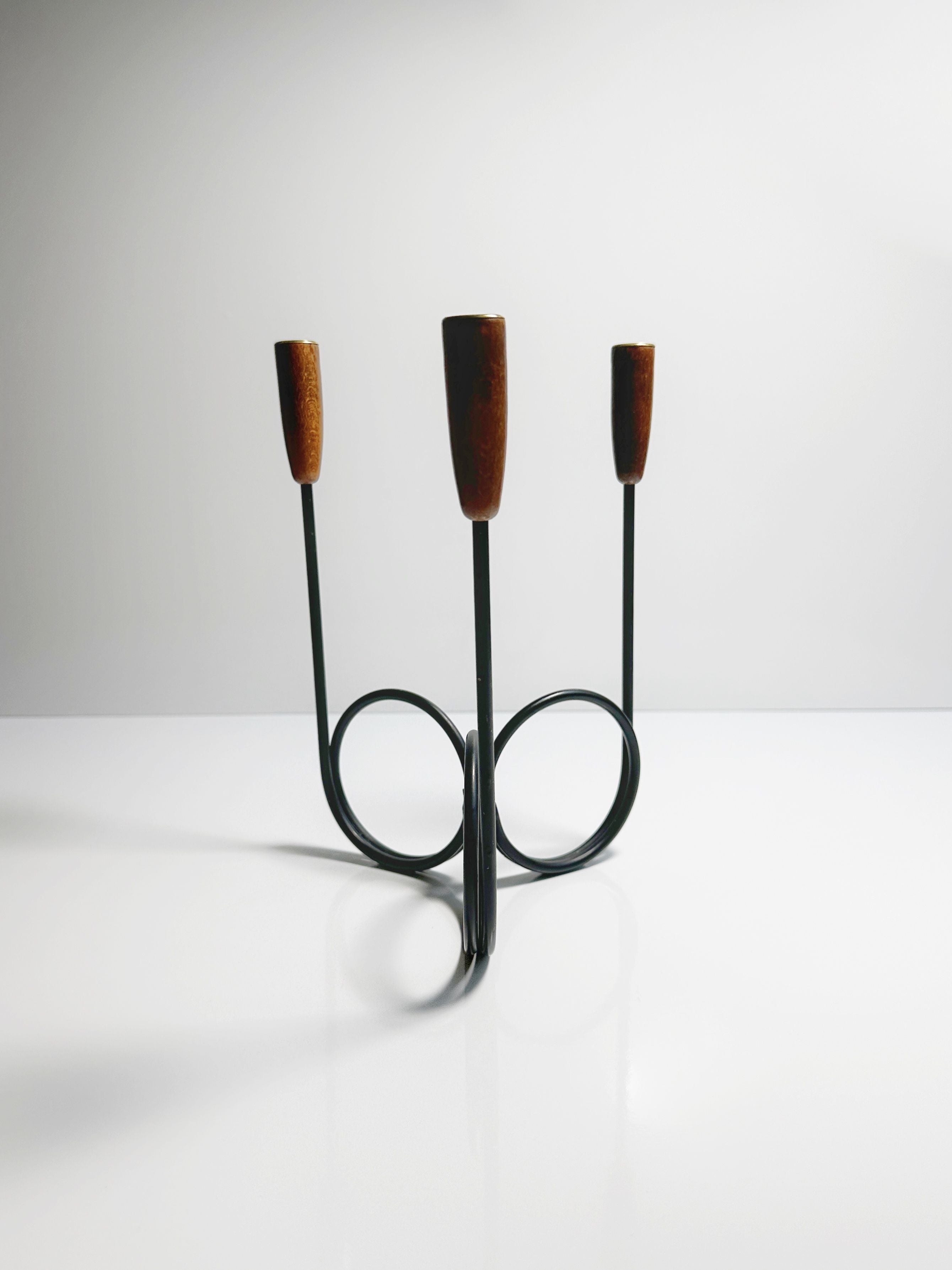 The Danish Candleholder 1960s by Dodo Vintage is a minimalist, modern three-armed candlestick holder featuring teak ends. Crafted from slender black metal rods that elegantly curve into a circular base, this stylish candelabra stands on a glossy white surface set against a plain light gray background.