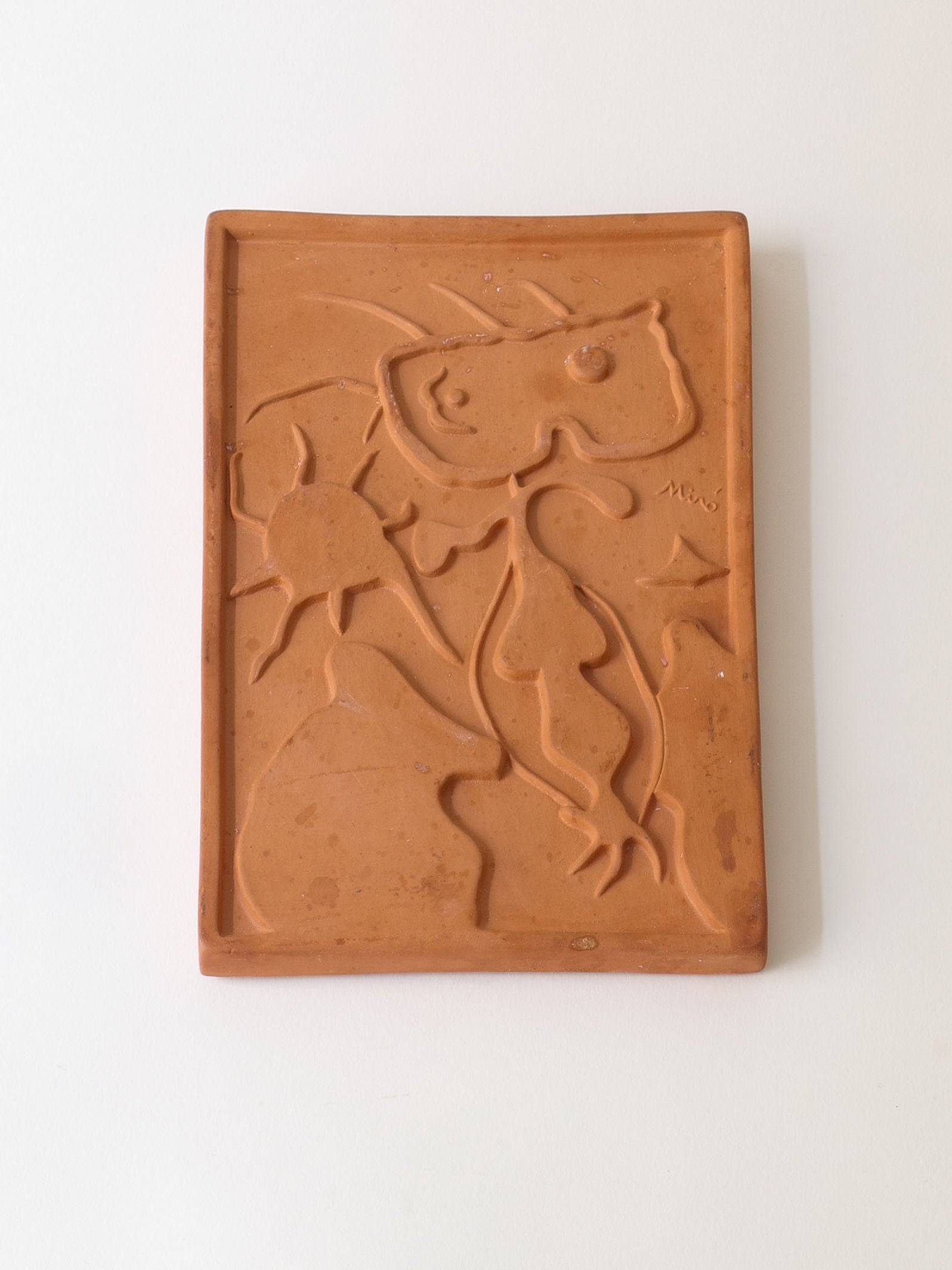 The Terracotta Relief Plaque by Collection apart, signed by Joan Miró (1938), features abstract embossed shapes and lines, a star, and organic forms, with "Miro" engraved on the right side against a plain white background, echoing the Catalan surrealist's style.