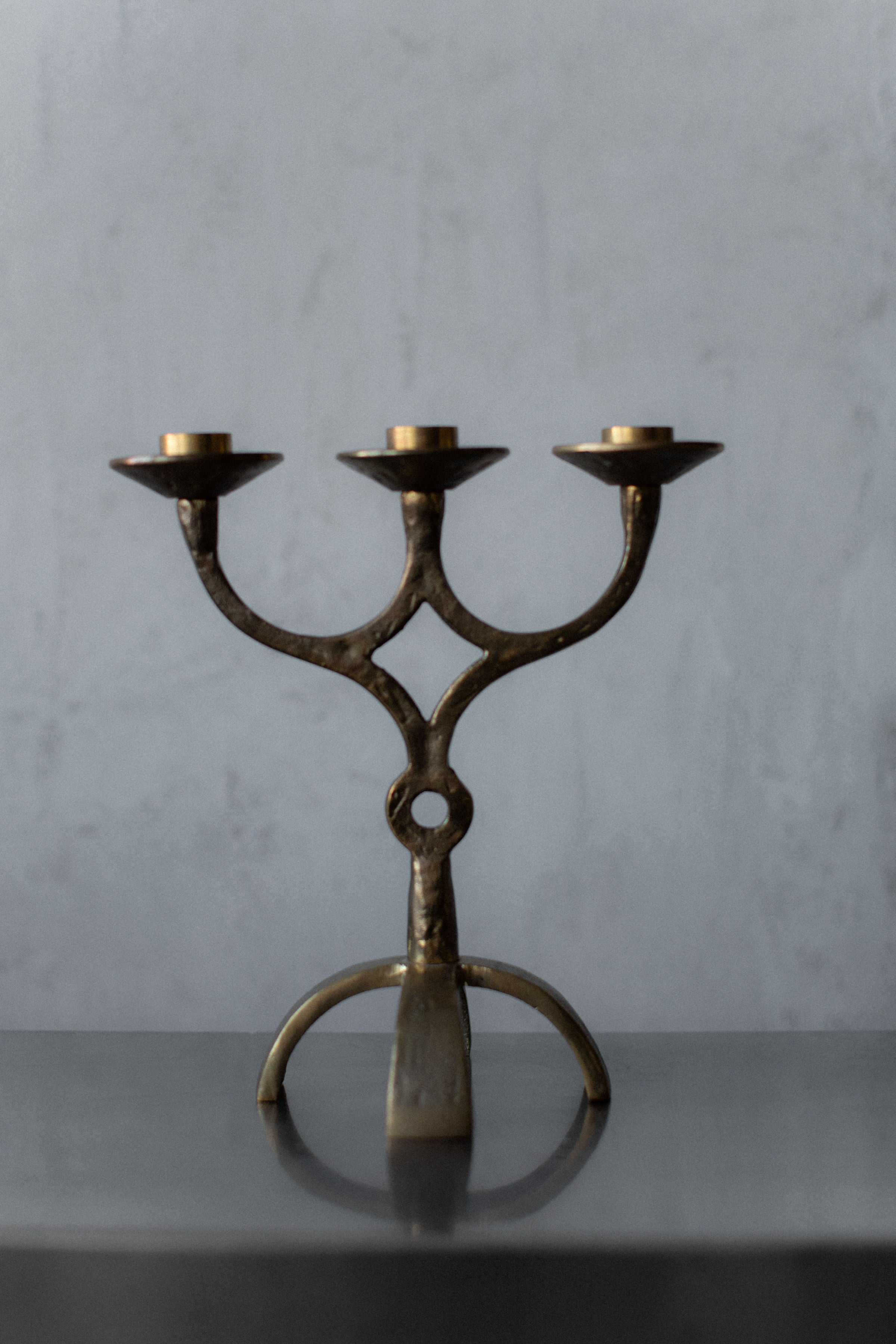 The Brutalist Brass Candleholder by Out For Lunch, a vintage three-armed centerpiece with intricate detailing, stands on a smooth dark surface against a muted gray wall. Each arm of the ornate design holds an unlit candle, showcasing its artisanal craftsmanship.