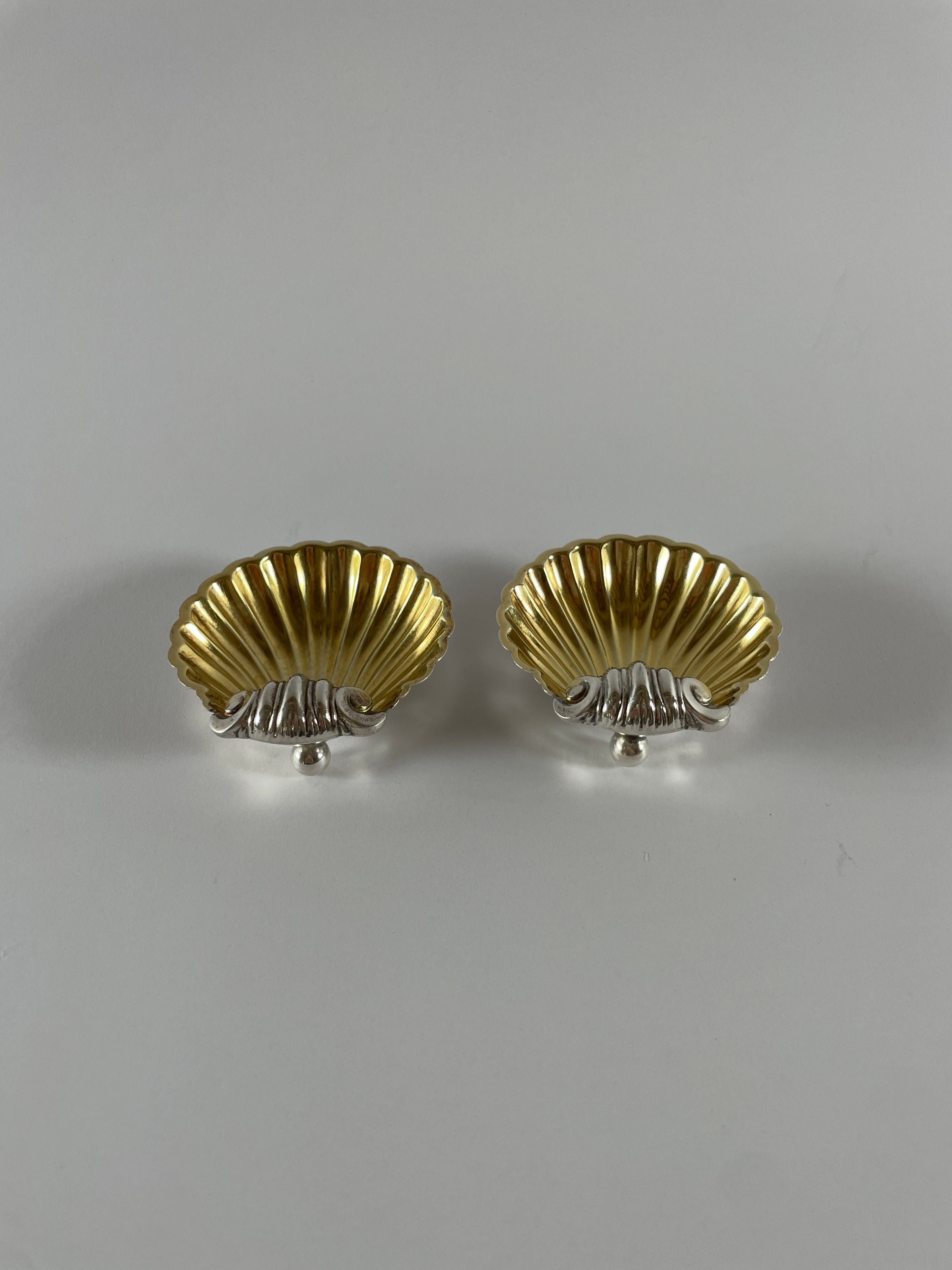 The "Vintage Silver Plate Shell Salts" by Maud Vaughan feature two elegant golden shell-shaped objects resting on a plain white background. Their ribbed texture and vintage silver plate bases are reminiscent of 1930s decor, positioned beautifully side by side.