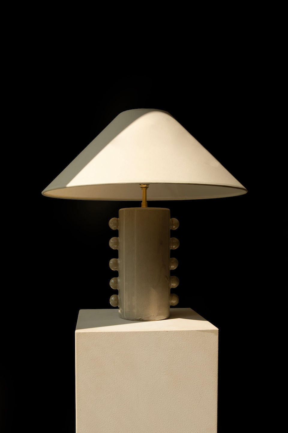 The 'Nicola' Table Lamp by Landy Rakoto Ceramics features a modern ceramic base adorned with pearl-like elements and a wide conic cotton shade. Displayed on a white pedestal against a black background, it highlights its elegant design.