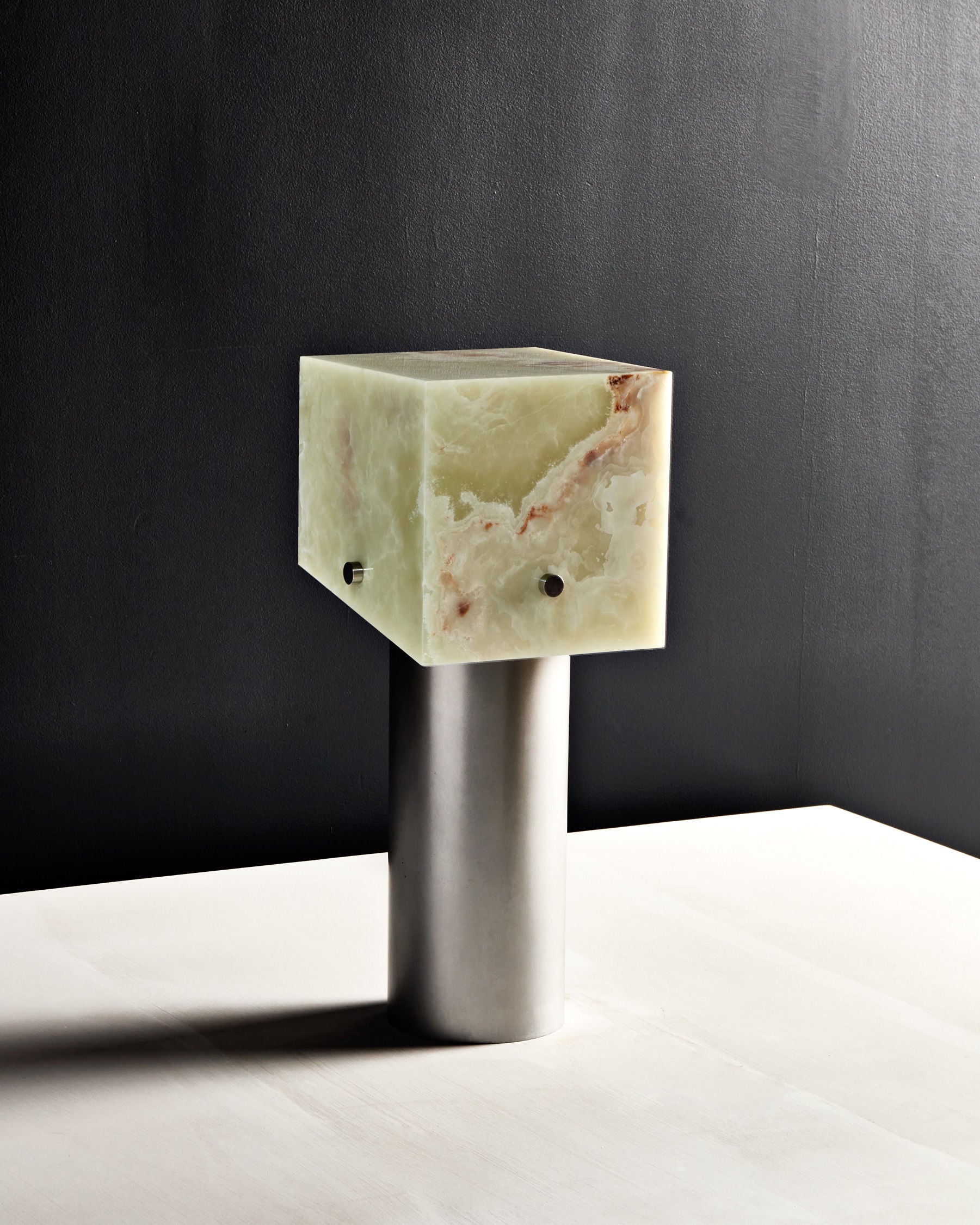 The 'Mada' Table Lamp by Slash Objects features a translucent green onyx-inspired cubic shade atop a sleek cylindrical metallic base, displayed on a white surface against a dark background.