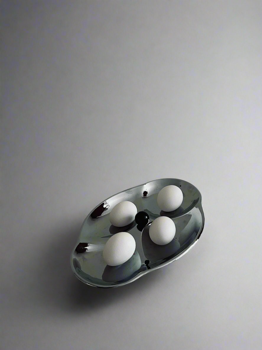 A sleek, modern Mid Century Serving Tray by Flock Studio, featuring an abstract design and a silver finish, holds four white eggs. Its reflective surface stands out against the plain, muted background, creating a minimalist and artistic composition reminiscent of Mid Century aesthetics.