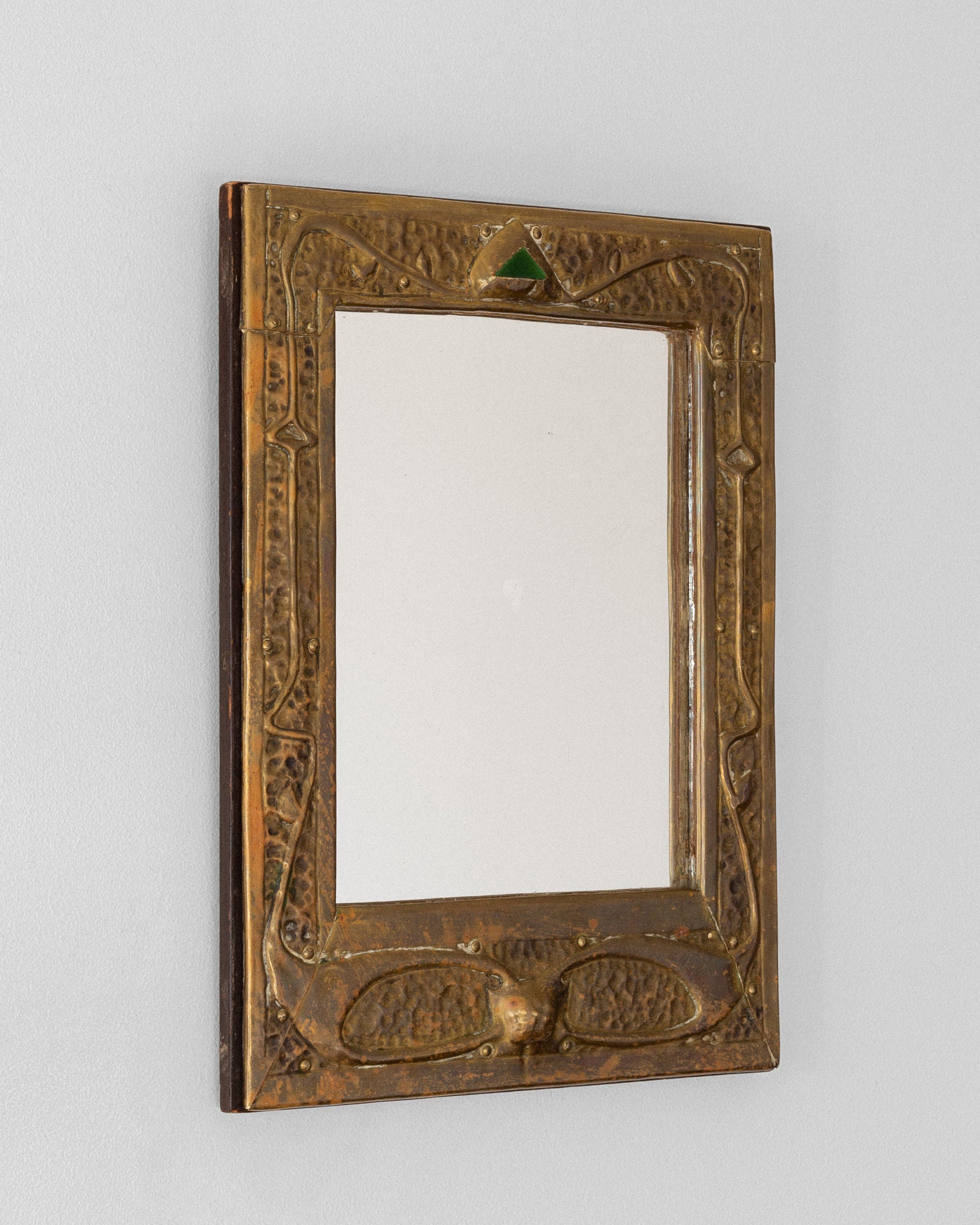 The Spigel Art Nouveau Brass Wall Mirror features an ornate hammered brass frame with an arched green jewel at the top, elegantly mounted on a plain light-colored wall.
