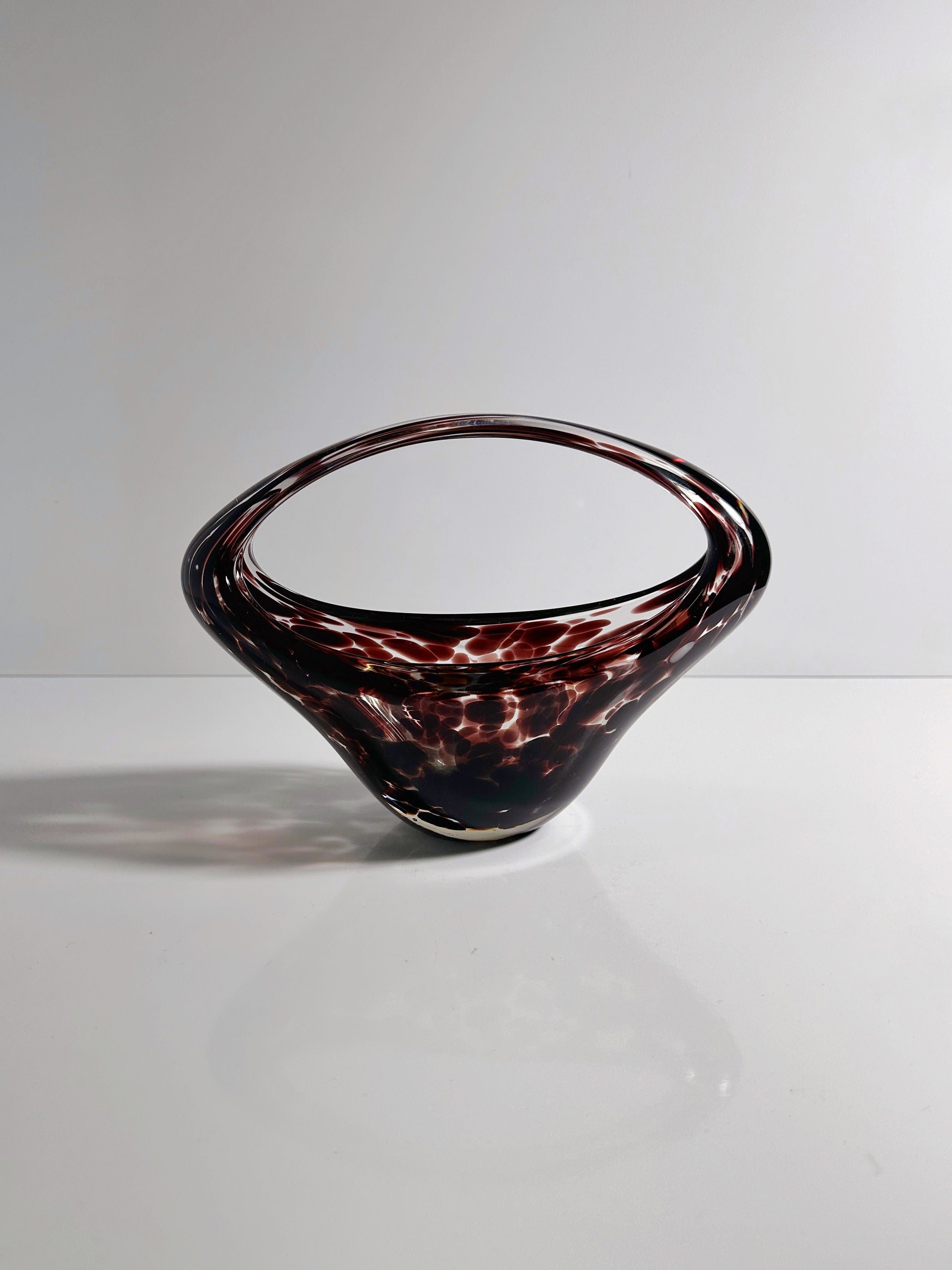 The Murano Tortoise Glass Handle Bowl by Dodo Vintage, a 1970s sculptural piece with dark swirling patterns, sits elegantly on a reflective surface. Its abstract, open curved design creates a sophisticated silhouette against the plain background.