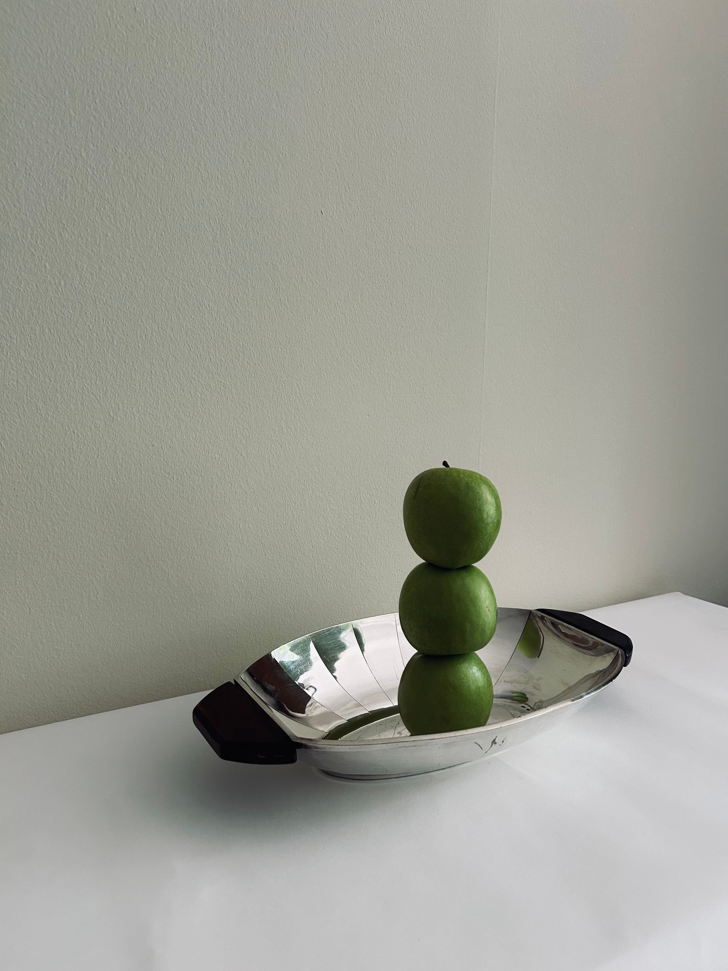 A minimalist image of three green apples stacked vertically on the Flock Studio Art Deco Serving Tray. The tray, crafted from polished metal with handles, rests on a white surface against a plain light-colored wall. Soft lighting casts subtle shadows, enhancing the modernist design's elegance.