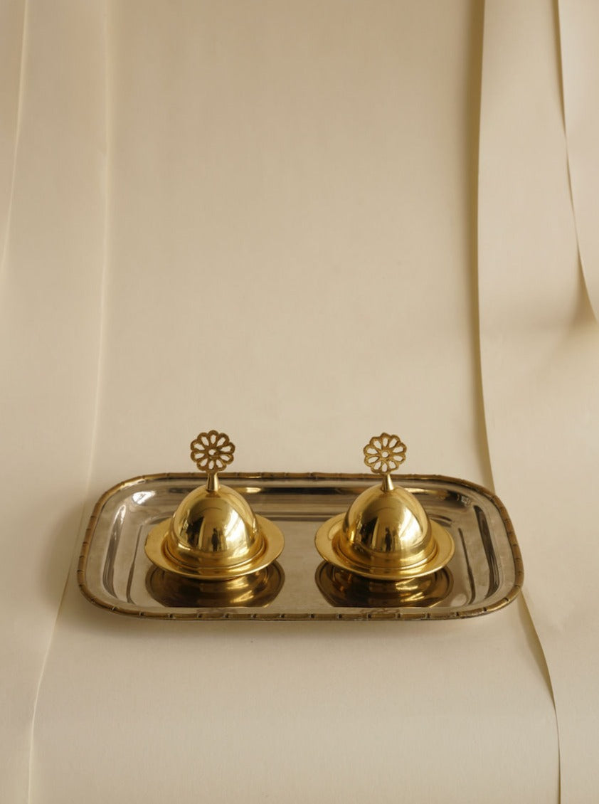A polished metal tray holds two ornate, golden domed containers with flower-shaped handles under intricate lids. The scene, featuring the flower tops against a neutral beige fabric draped in soft folds, exudes elegance and simplicity. This exquisite arrangement features the Art Nouveau Plate With Dishes by Les Objets Amsterdam.