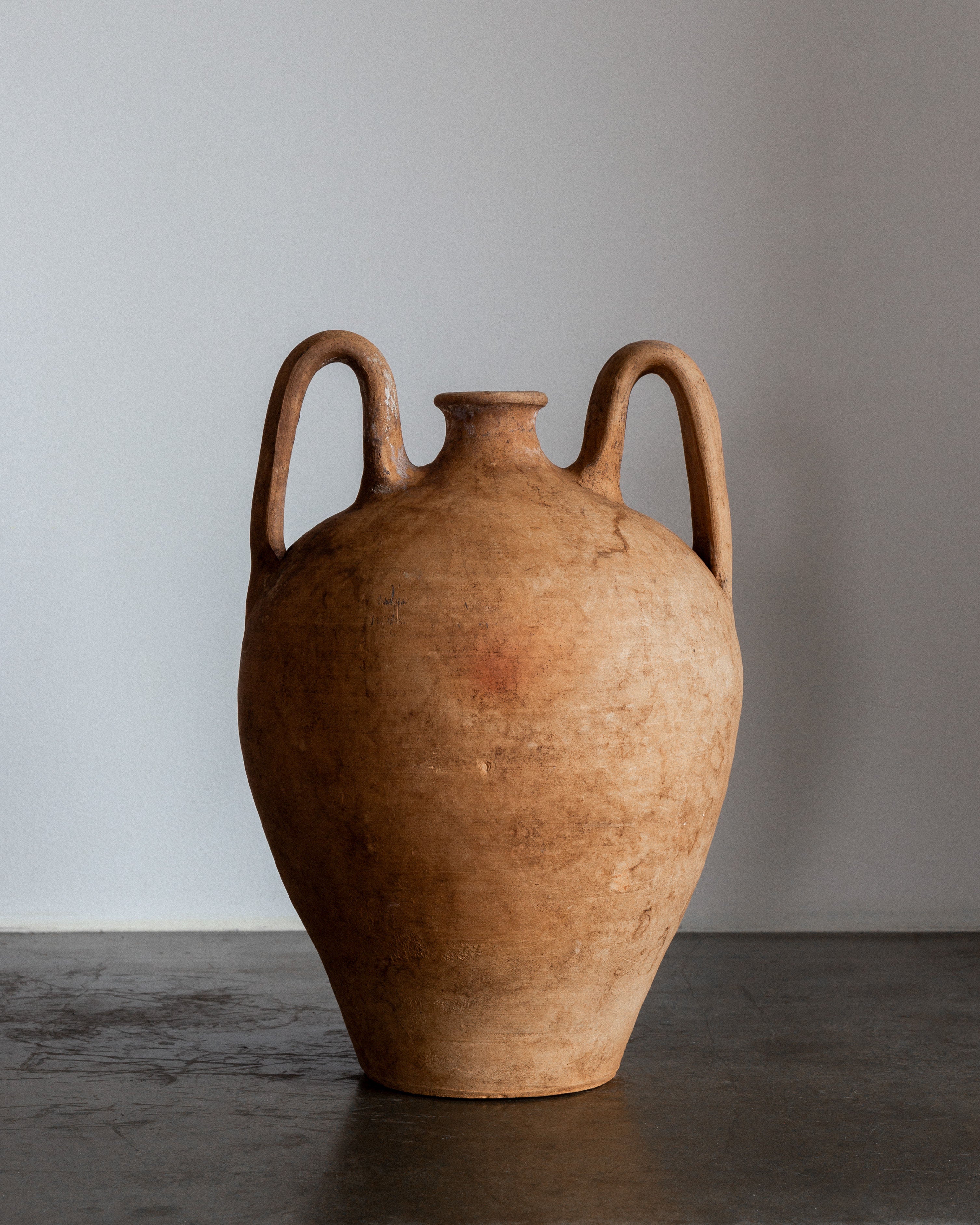 A stunning decorative piece, the Spigel Early 20th Century European Ceramic Vase features a weathered texture, earthy tones, wide body, narrow neck, and two curved handles. It stands on a dark surface against a plain gray background.