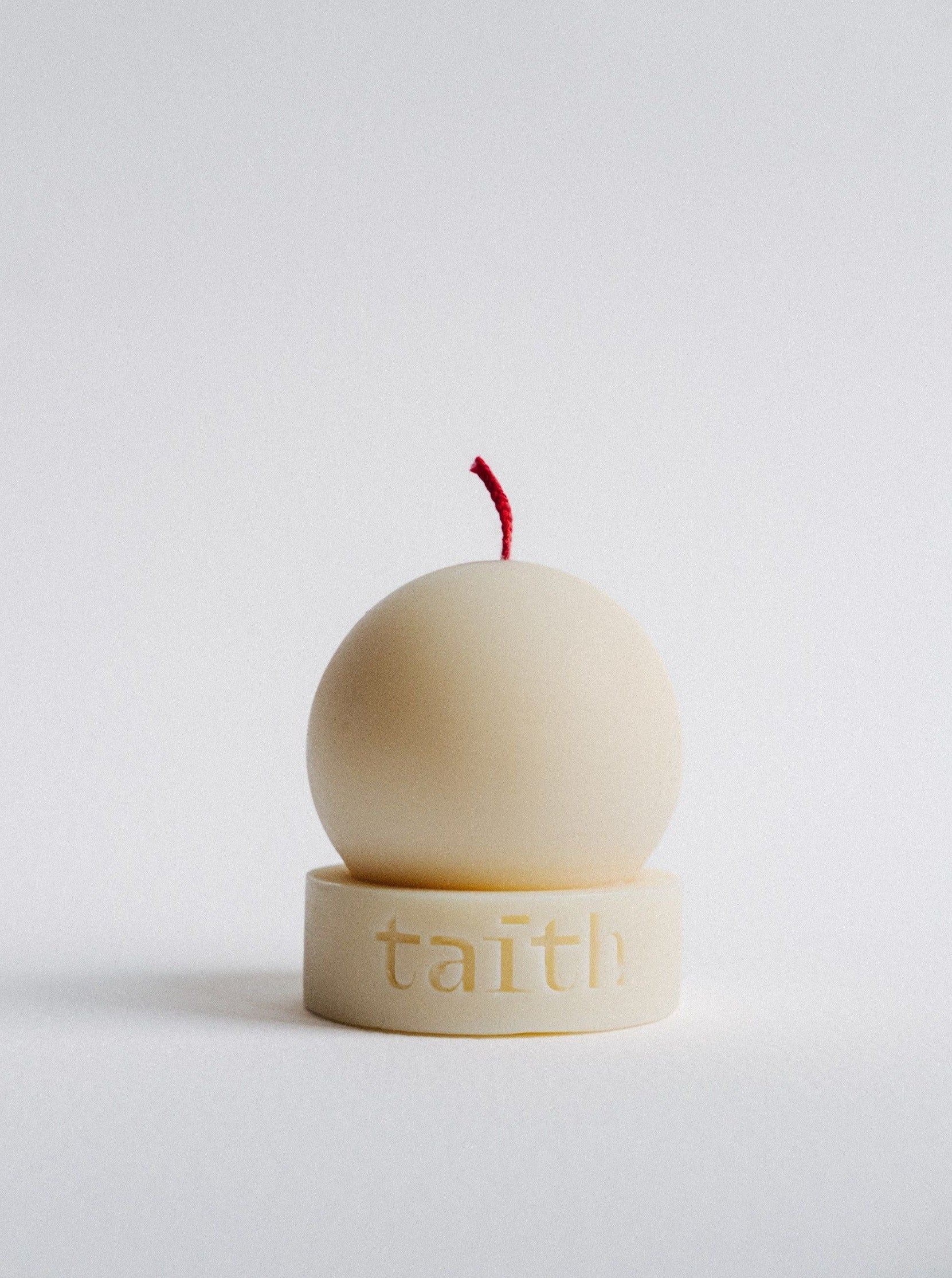 A round, beige-colored Taith Lu Candle Cream with a red wick sits on a matching circular base engraved with the word "taith" in lowercase letters against a plain white background. The minimalist design and captivating fragrance give the candle a clean and simple aesthetic.