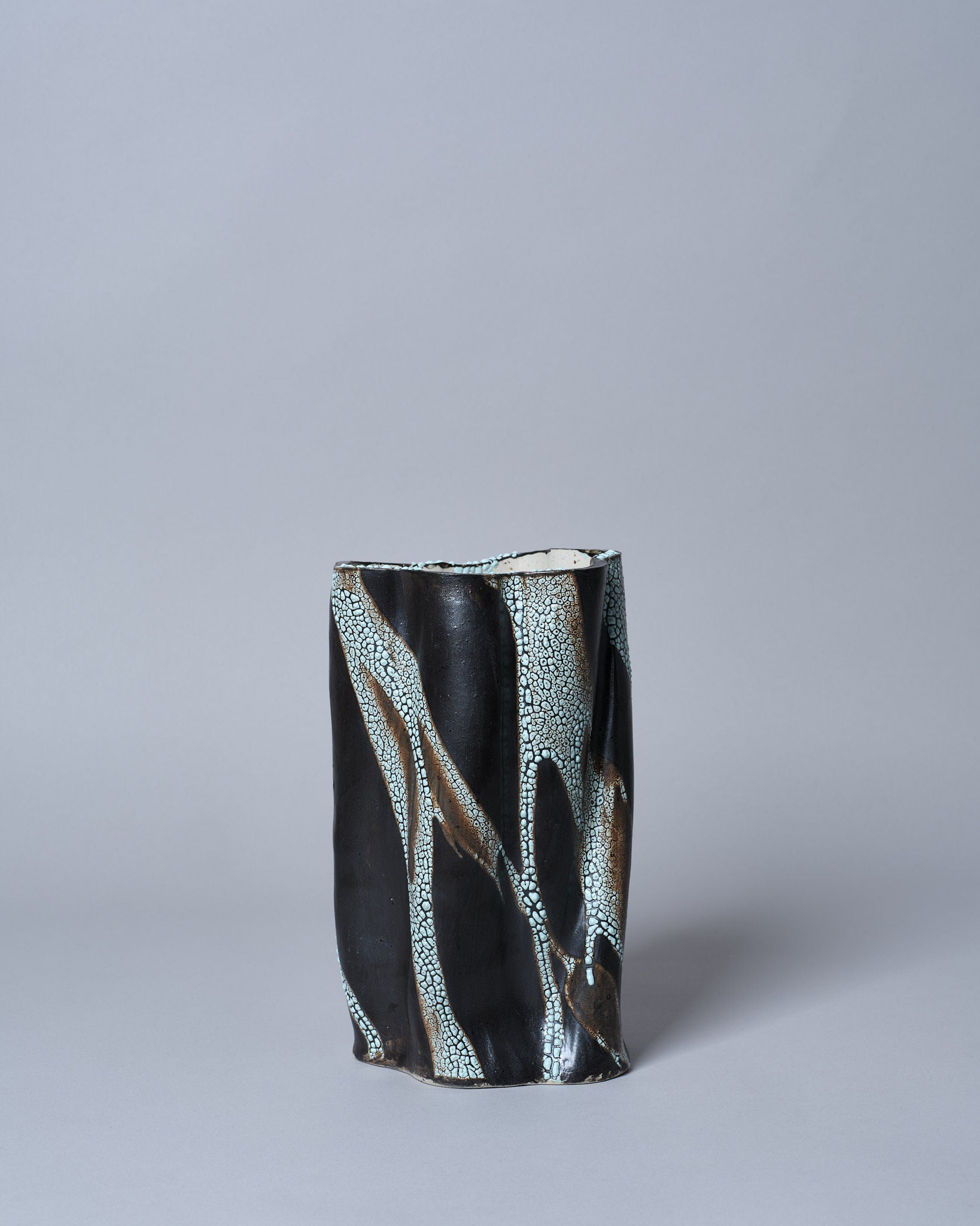 The Swirly Vase Brown and Light Blue by Astrid Öhman is a tall, uniquely-shaped stoneware vase with a wavy edge, featuring a dark, glossy finish with textured white and light brown crackle patterns on a plain gray backdrop. This handmade piece adds an artistic touch to any setting.