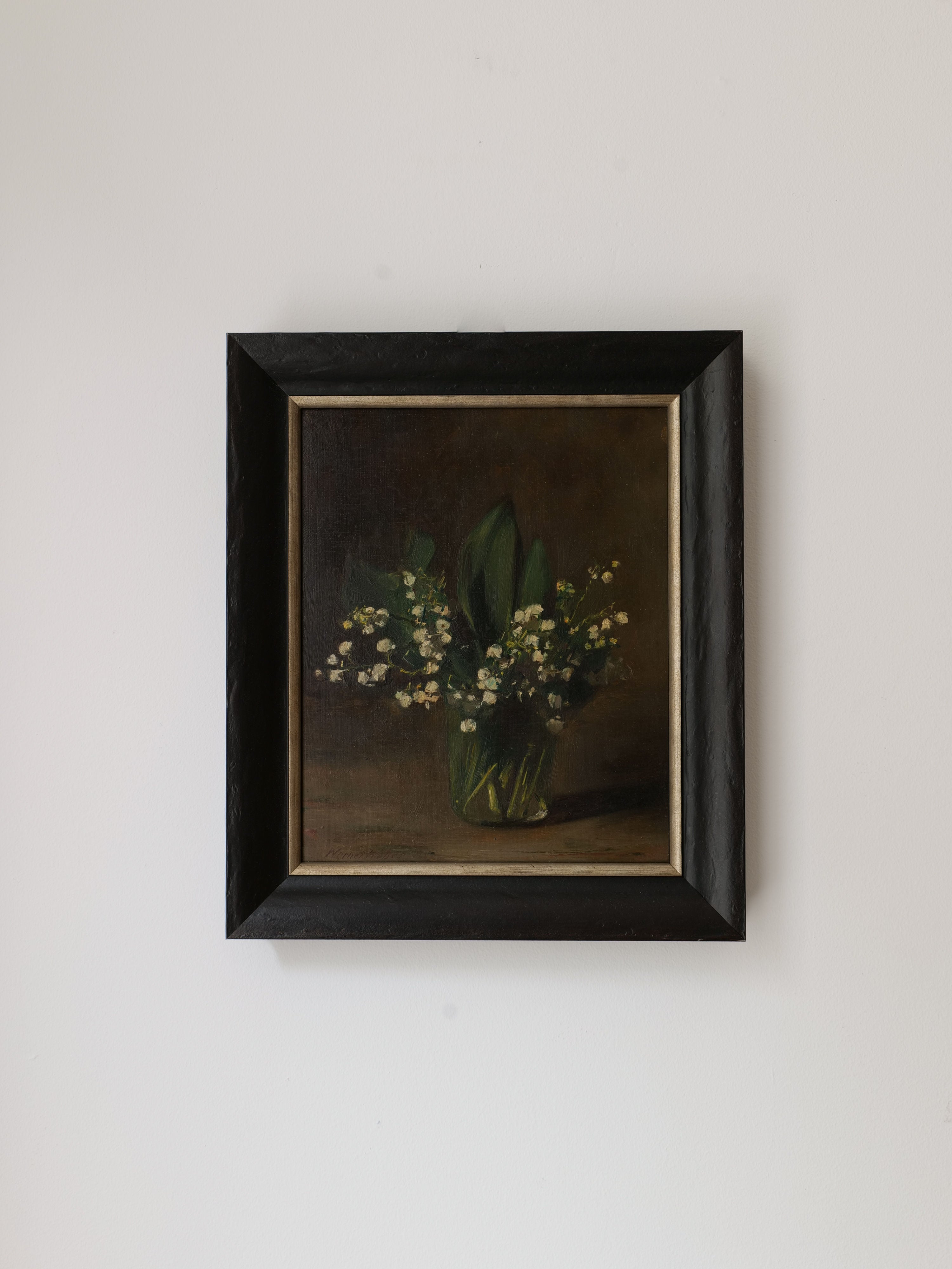 A framed painting, Lily of the Valley Still Life 1961 by Collection apart, of a green vase filled with small white flowers and green leaves, reminiscent of a classic Dutch still life, graces the plain white wall. The simple black frame contrasts beautifully with the painting's dark tones.