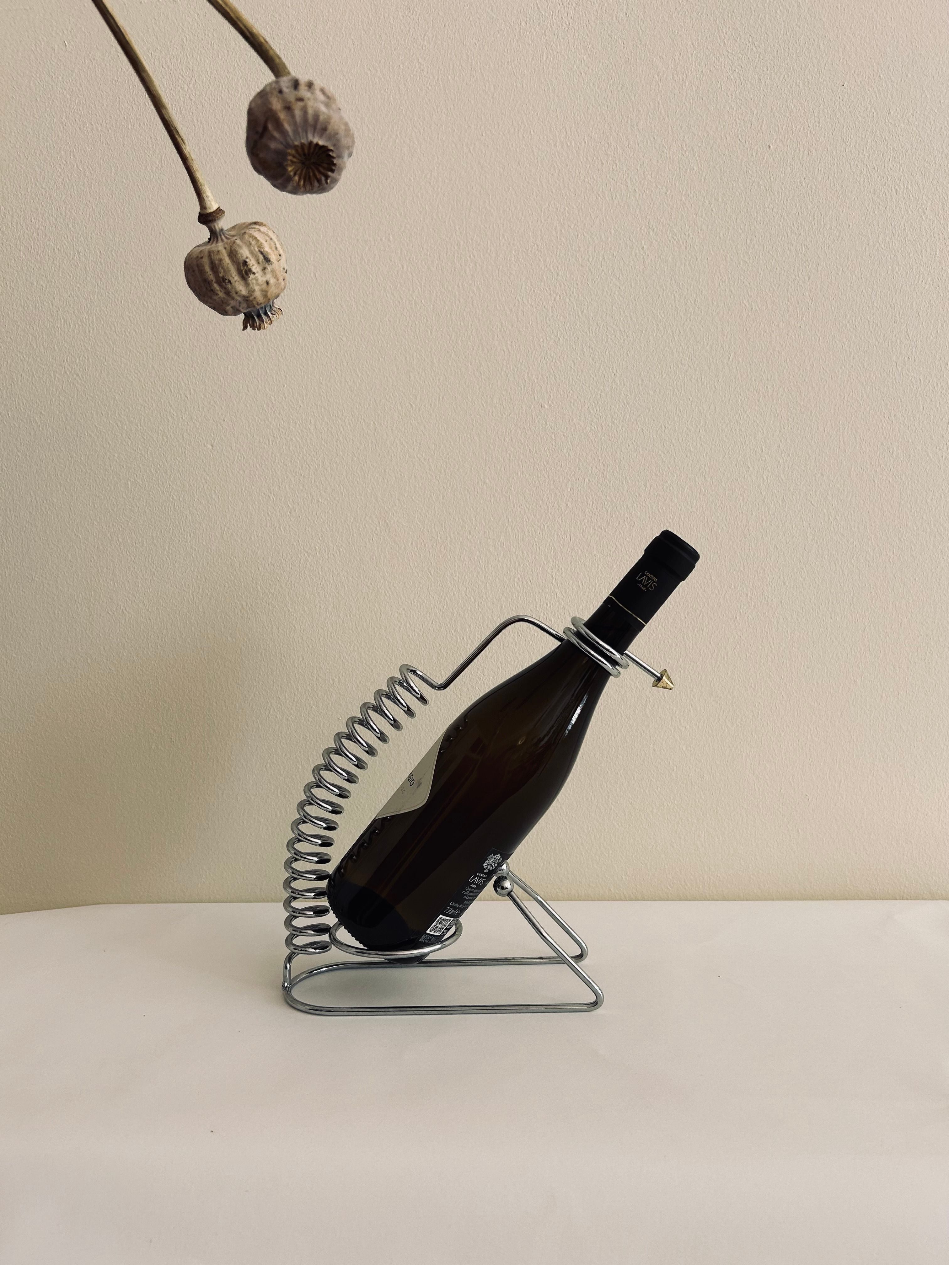 The Postmodern Wine Holder by Flock Studio cradles a wine bottle in its sleek metal spiral on a light surface, enhancing the elegant ambiance. Dried poppy pods hang in the upper left corner against a plain beige background, perfecting the minimalist aesthetic.