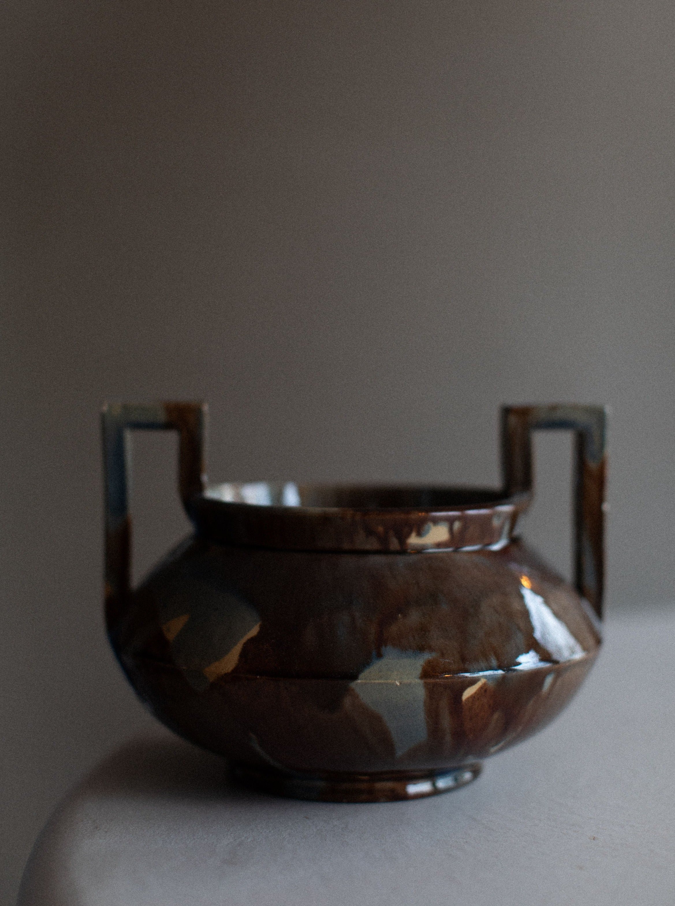 An ancient ceramic bowl with handles sits on a surface, featuring a glossy finish with cracked and chipped sections, illuminated by soft, natural light. Its style echoes the vintage sophistication of traditional Swedish design. The 1930's Höganäs Vase by Out For Lunch.