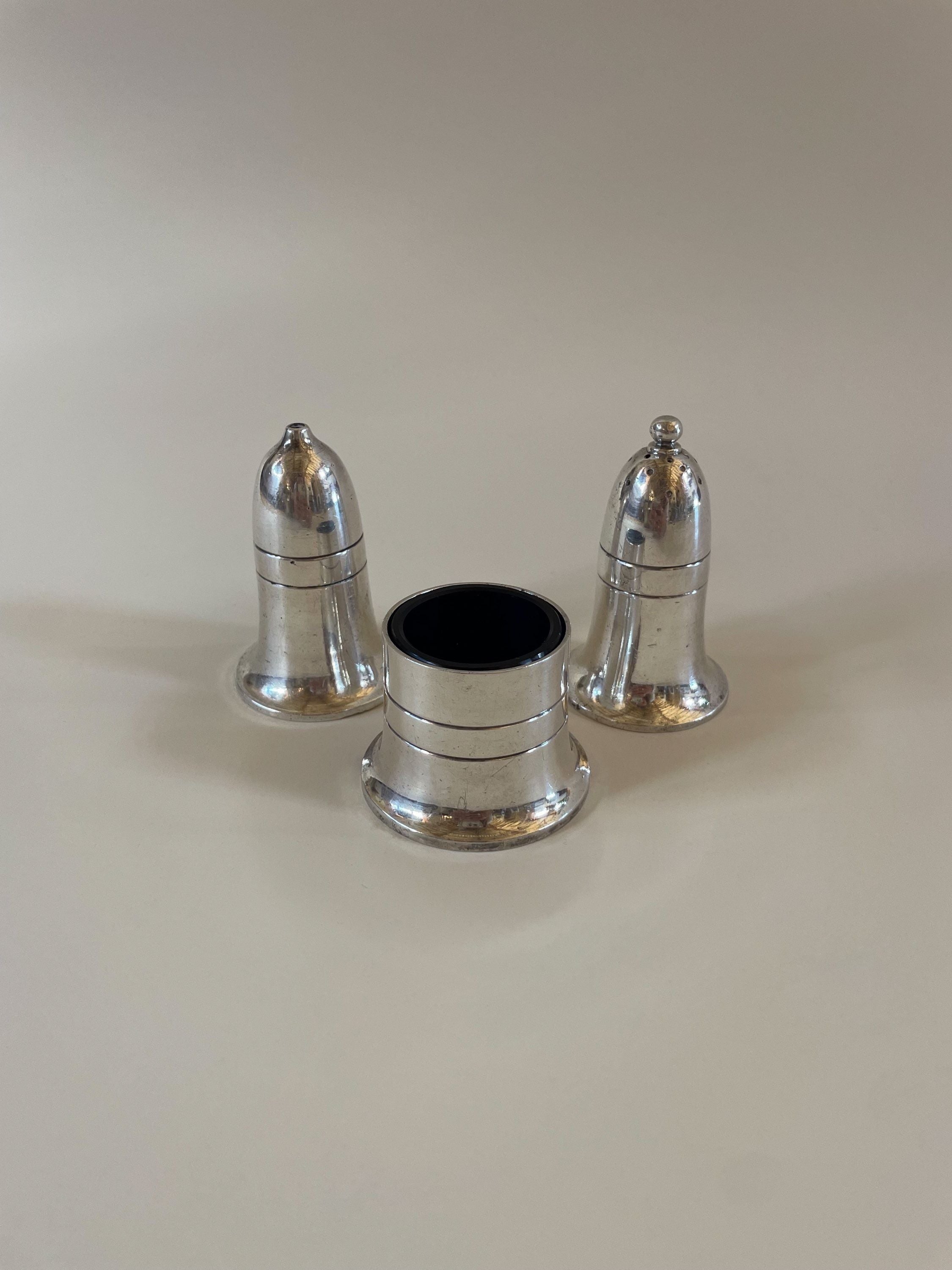 Three small silver-colored metal objects are arranged on a flat surface. Two objects resemble traditional bell-shaped pepper shakers, while the third is shorter and cylindrical with an open top, possibly a napkin ring or holder. This setup evokes the charm of a Vintage Mappin and Webb Cruet Set by Maud Vaughan.
