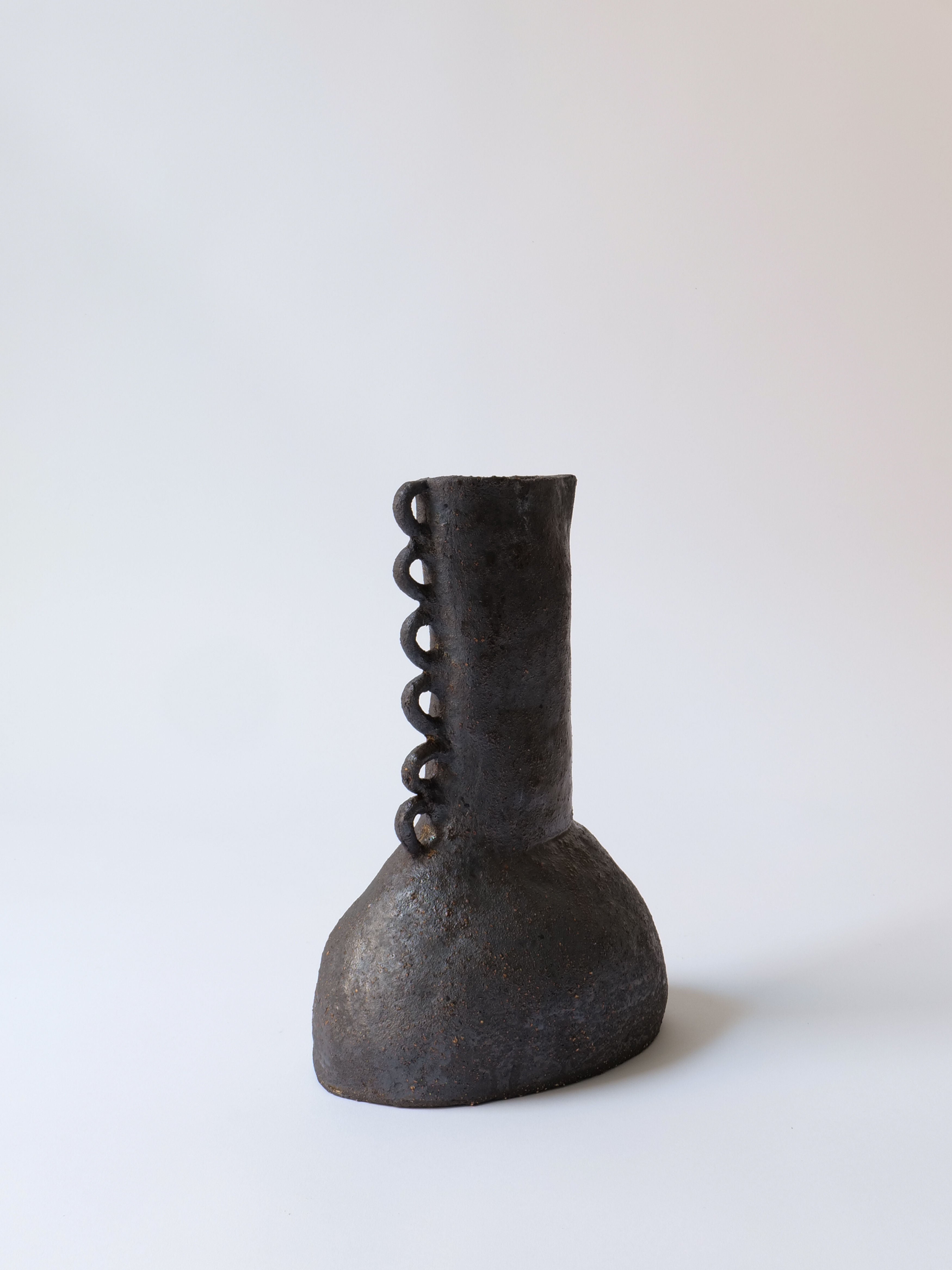 The Dragon Vase Oikos Estudio, crafted by Collection apart, embodies the Wabi-Sabi aesthetic with its uniquely shaped stoneware ceramic design. It features a rough texture, a bulbous base, and a tall, narrow neck adorned with circular looped decorations along one side. Displayed against a plain light background, this black vase exudes subtle elegance.