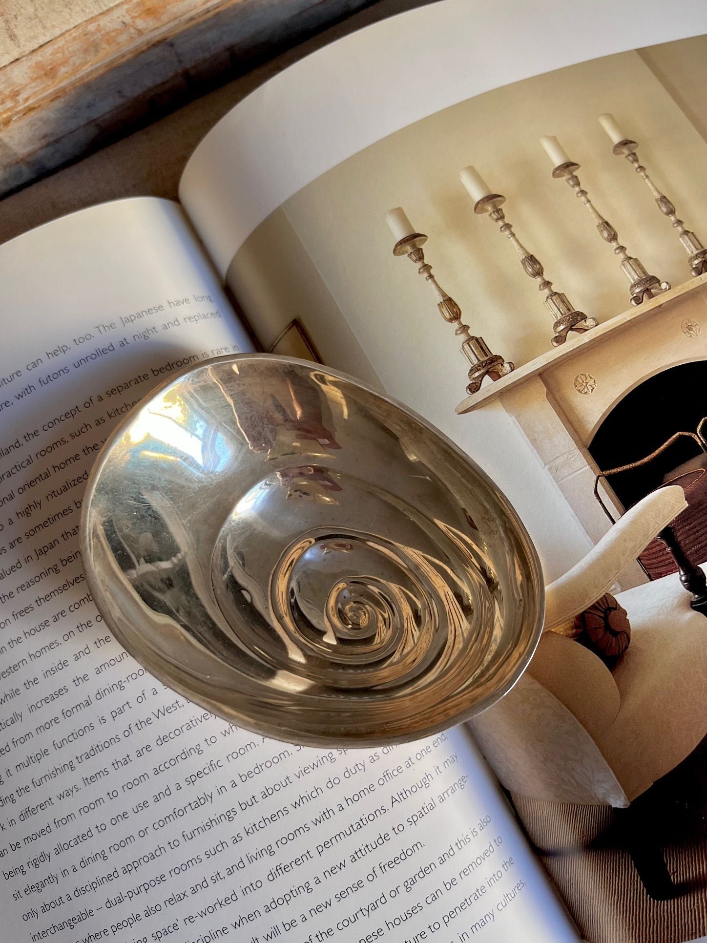 A stunning Rare Sterling Silver Midcentury Bowl by Médecine lies on an open book, its pages revealing text on the left and an interior photograph featuring a midcentury fireplace with four candlesticks on the right. The edge of a white couch is visible beside the welcoming fireplace.