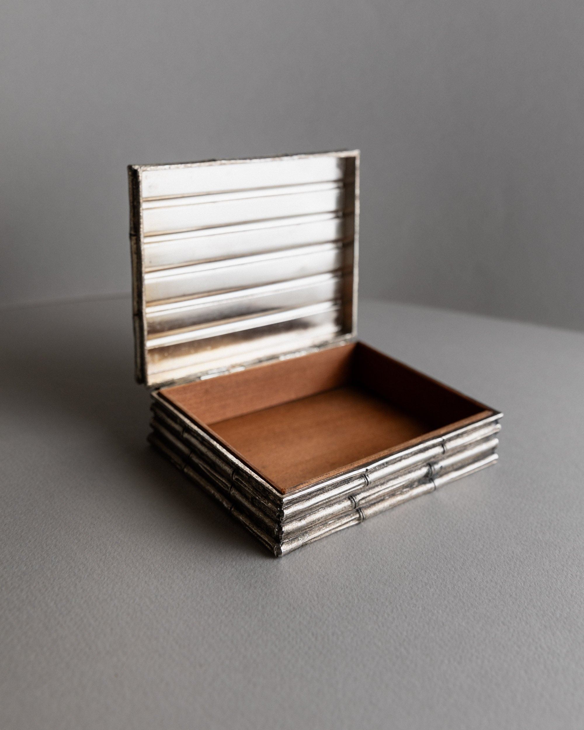 A Bottega Jacobs Box in Silver-plated from 1970s France, with a ribbed exterior and plain wooden interior, rests on a gray surface. Its hinged lid stands upright, capturing a retro chic vibe that seamlessly merges classic elegance with modern style against the matching backdrop.
