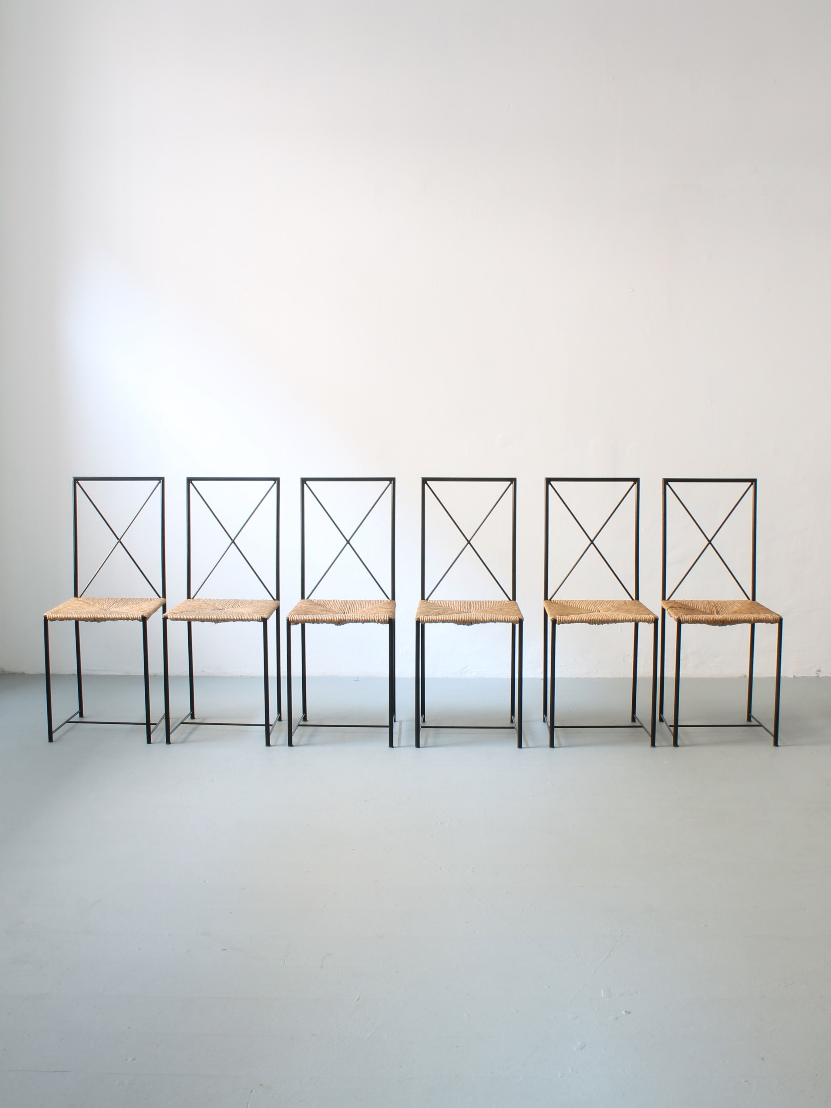 Six "Moka" chairs by Mario Asnago & Claudio Vender for Afterhour Furniture, featuring plant fiber seating and an open-back "X" design in lacquered tubular metal, are lined up against a plain white wall on a light gray floor.