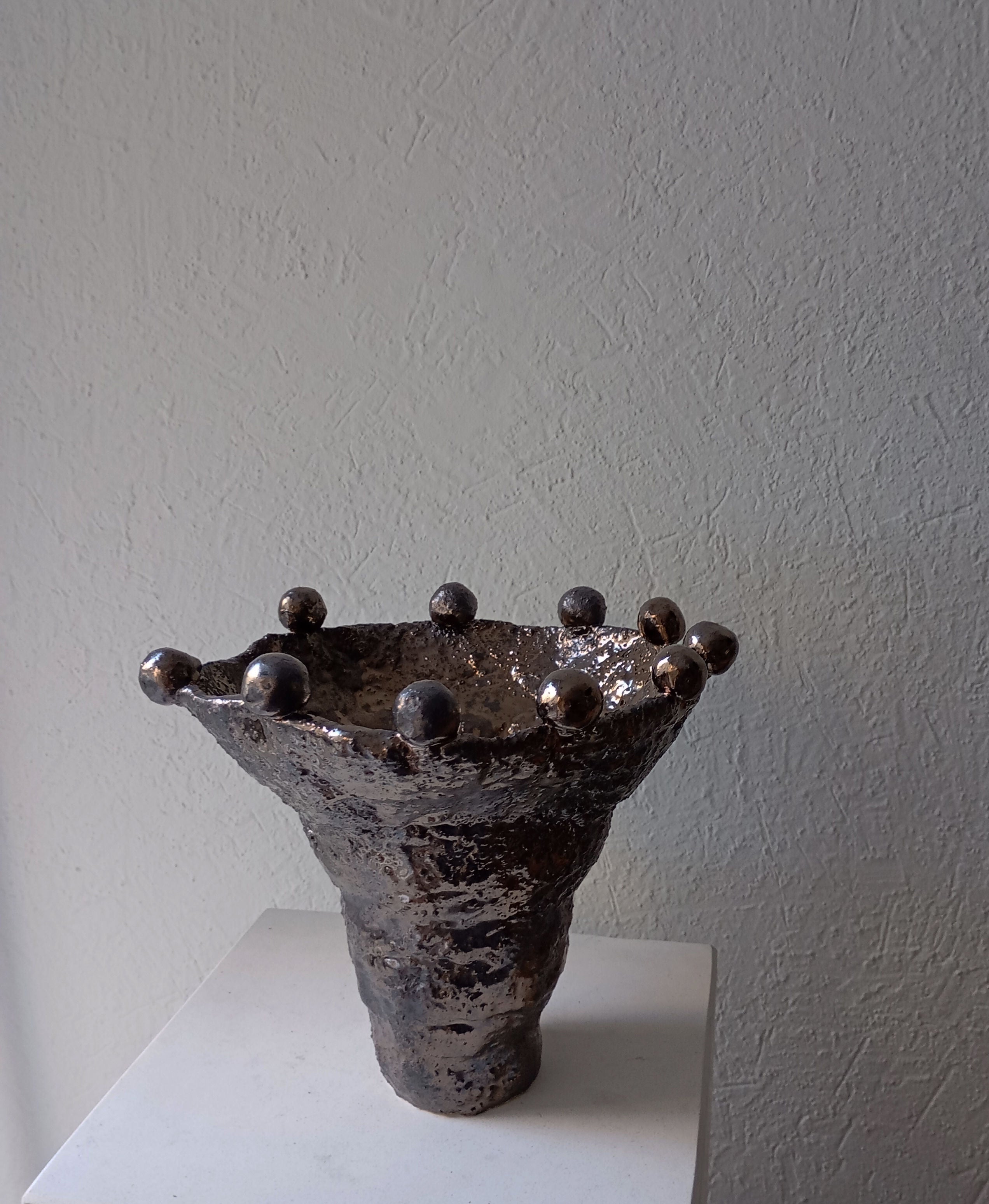 The Coupe Lilibeth 3 by Claire Pain is a unique metallic textured ceramic vase with a flared rim and rounded protrusions, displayed on a white pedestal against a light textured wall, evoking sculptural bronze enamel artistry.