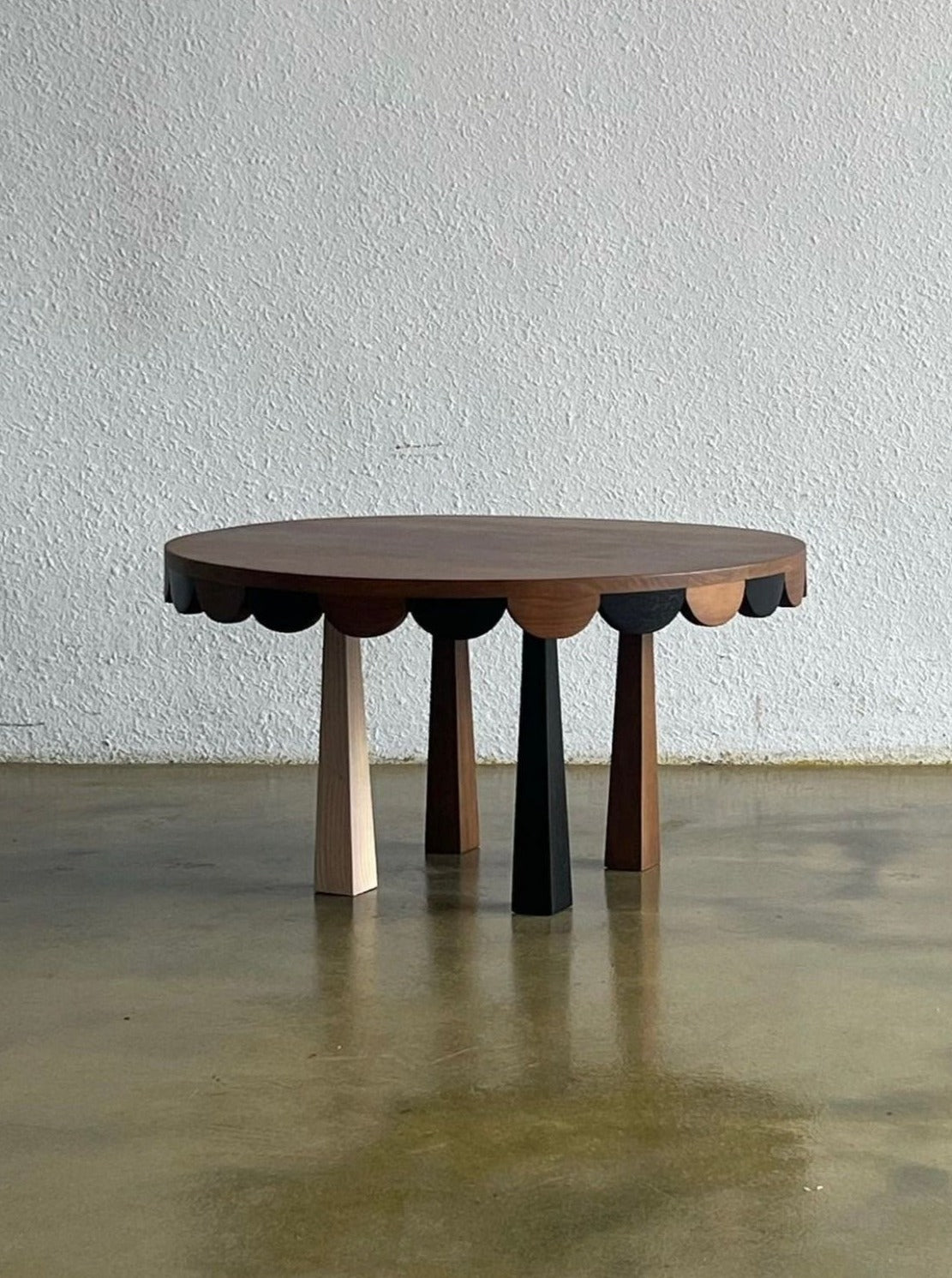 A round, handcrafted solid wood coffee table with a scalloped edge design, featuring four multicolored, conical legs in shades of black, dark brown, light brown, and beige. Made from teak wood, the Tent Coffee Table by Studio Kallang is placed on a smooth, reflective concrete floor against a textured, light-grey wall.
