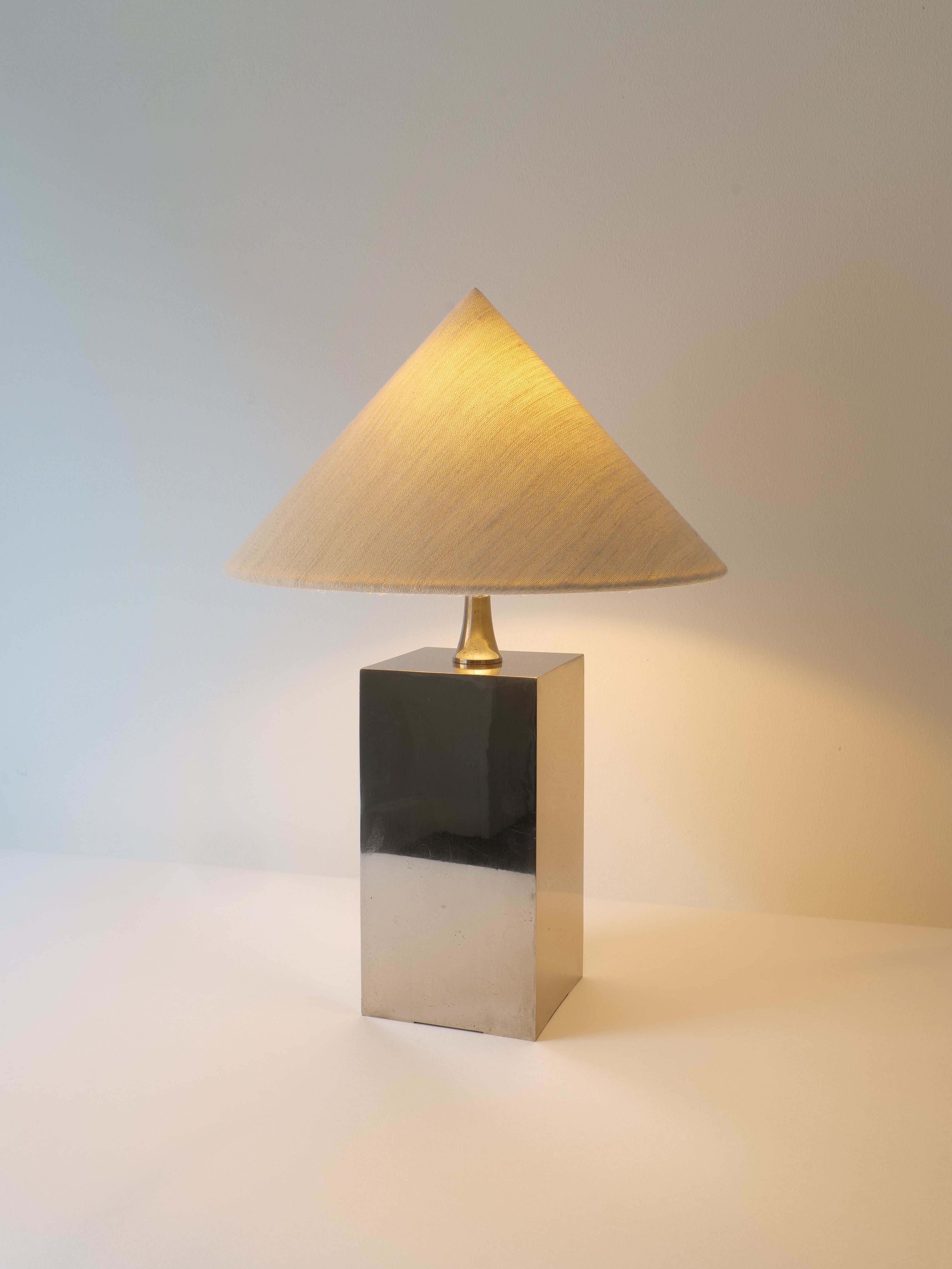 A modern Barbier Small Box Lamp 1970s by Collection apart with a rectangular chrome over brass base and a conical fabric lampshade is illuminated, casting a warm glow. The French 1970s lighting piece is positioned against a plain, light-colored background.
