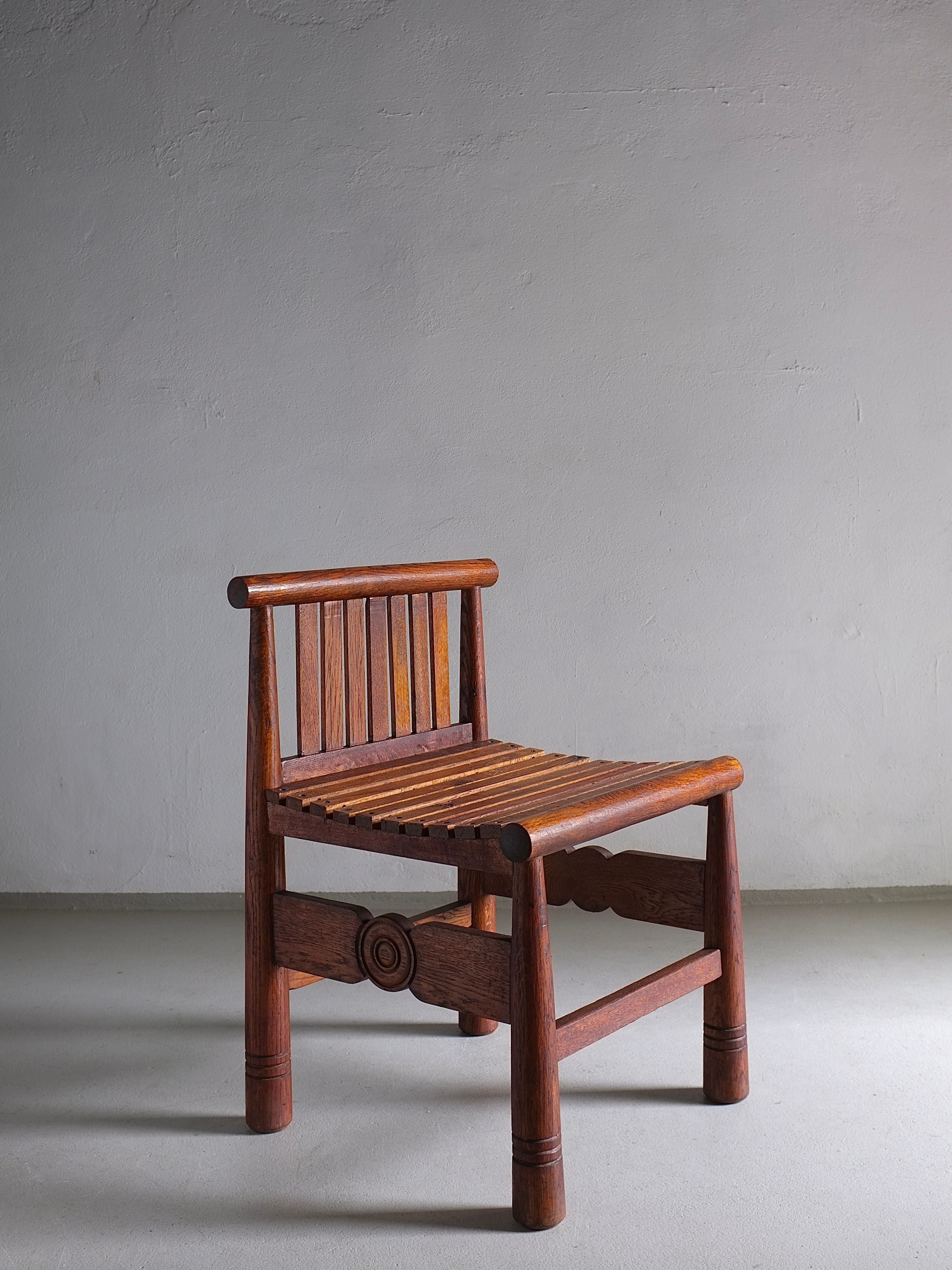 A Carved Oak Low Back Chair by Veter Vintage, dating back to the 1960s in France, sits against a plain, greyish wall. The chair features a simple, rustic design with straight vertical slats on the backrest, rounded legs, and a distinctive carved circular pattern on the front stretcher. Its dark-stained oak finish exudes a rich and warm ambiance.