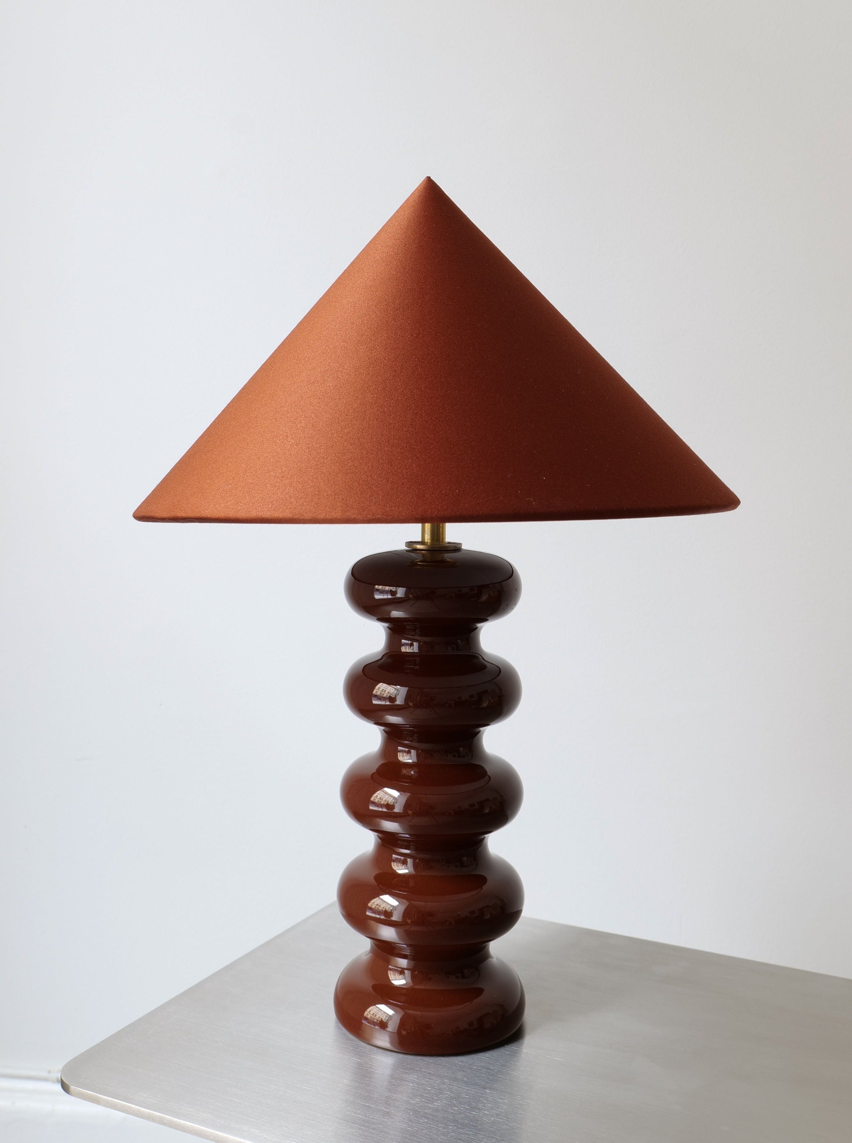 A handmade lampshade crowns this Sculptural Ceramic Lamp by Collection apart with a unique, wavy brown base, displayed against a plain white background. The large, brown cone-shaped fabric shade complements the lamp beautifully as it rests on a smooth, metallic surface.