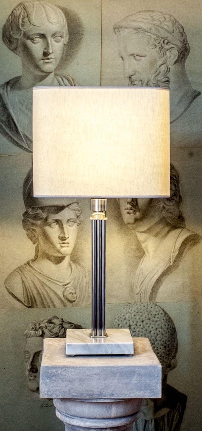 The Athens Table Lamp 3 by Brajak Vitberg, featuring a modern rectangular shade on a classic pedestal, is elegantly placed before an artful wall showcasing ancient Greek busts and sculptures, enriching the luxury interior with historical ambiance and sculptural sophistication.
