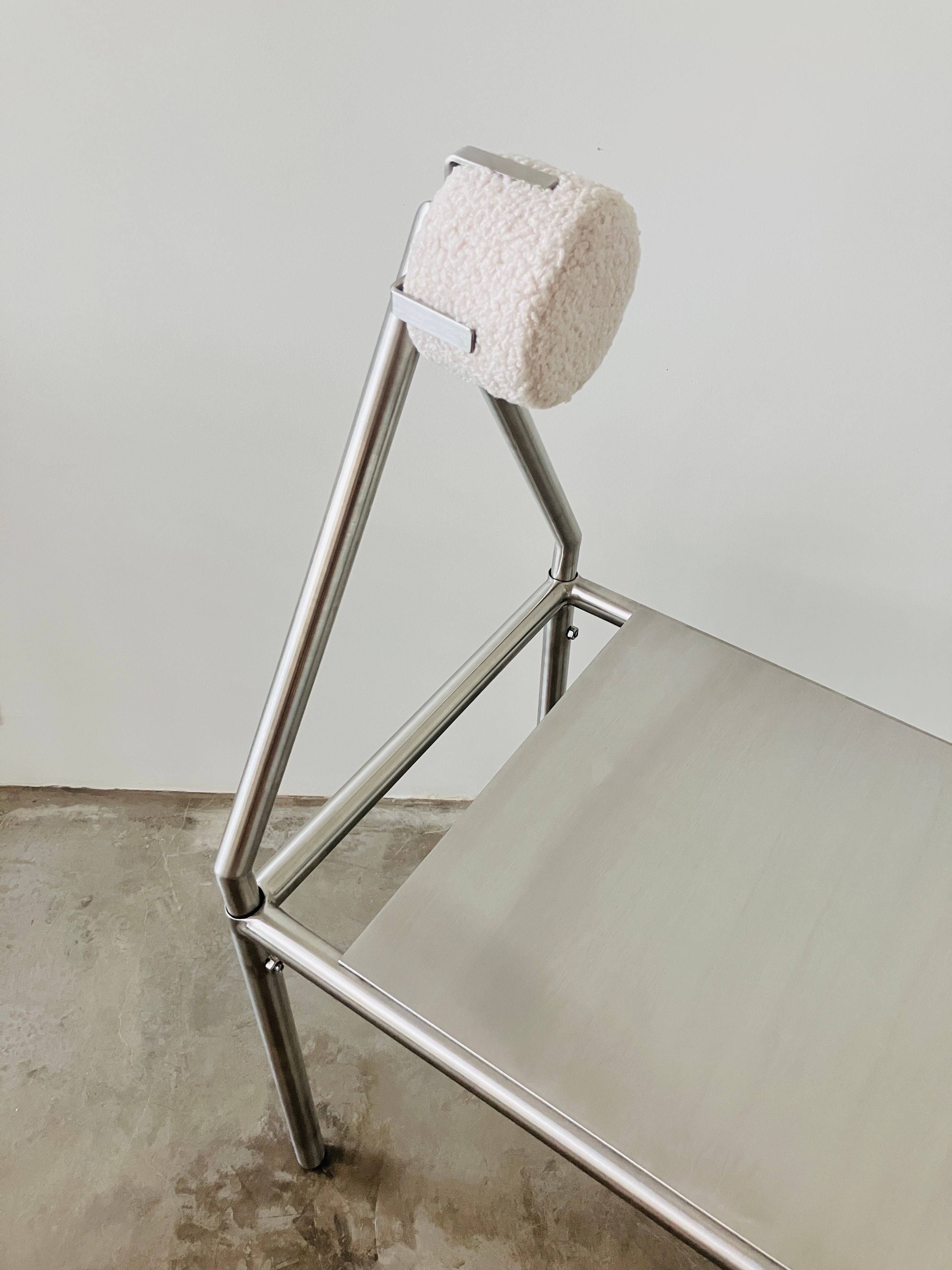 The 'H01' Stainless Steel Chair by HT features a minimalist metal frame, a flat seat, and a cylindrical textured white cushion atop the backrest. It’s shown on a concrete floor against a white wall.