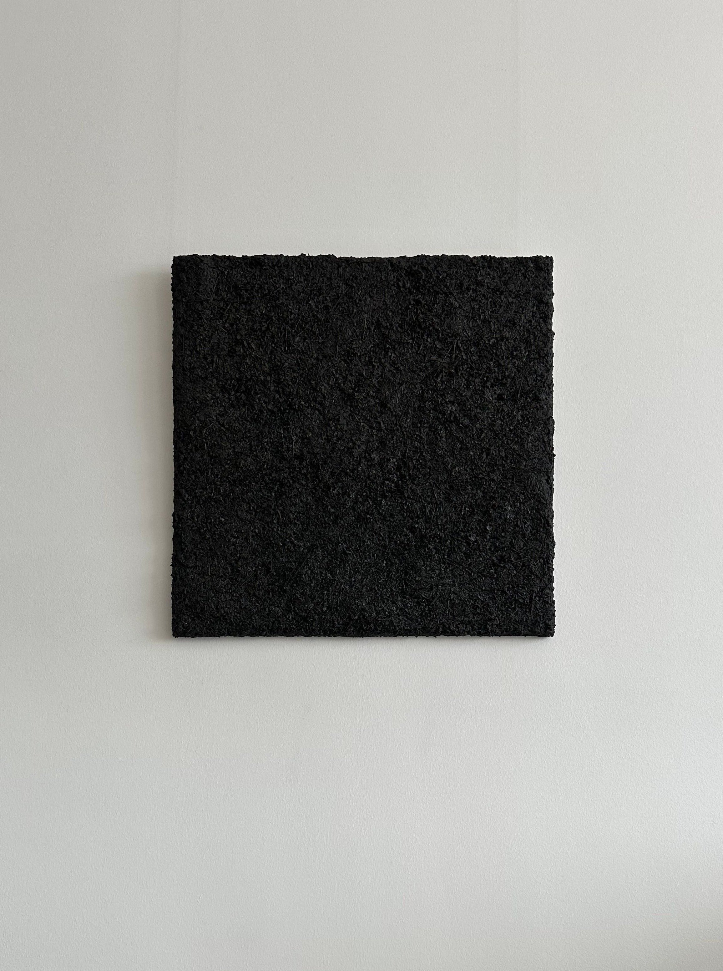 A minimalist piece of mixed media on canvas featuring a square with a rough, textured black surface, mounted on a plain, light gray wall. This made-to-order artwork, ARCHE 01 - Mixed media on canvas by CARRARA Studio, is simple and monochromatic, offering unique flair to any space.