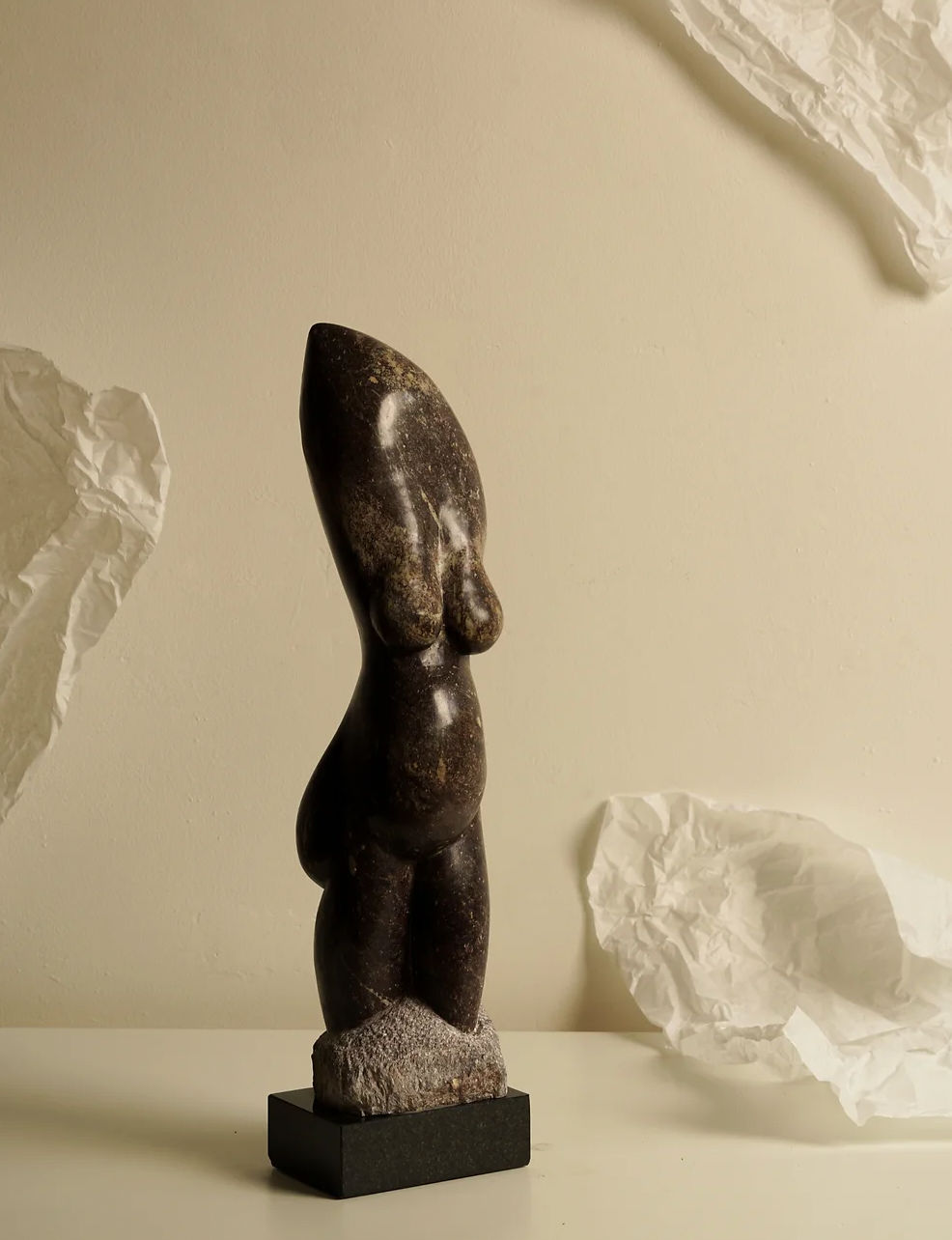 The Marble Femininity Sculpture by Les Objets Amsterdam features a curvy, abstract representation of the female form in dark marble, set on a black base. It is enveloped by crumpled white paper against a neutral backdrop, offering an artistic and minimalist aesthetic.