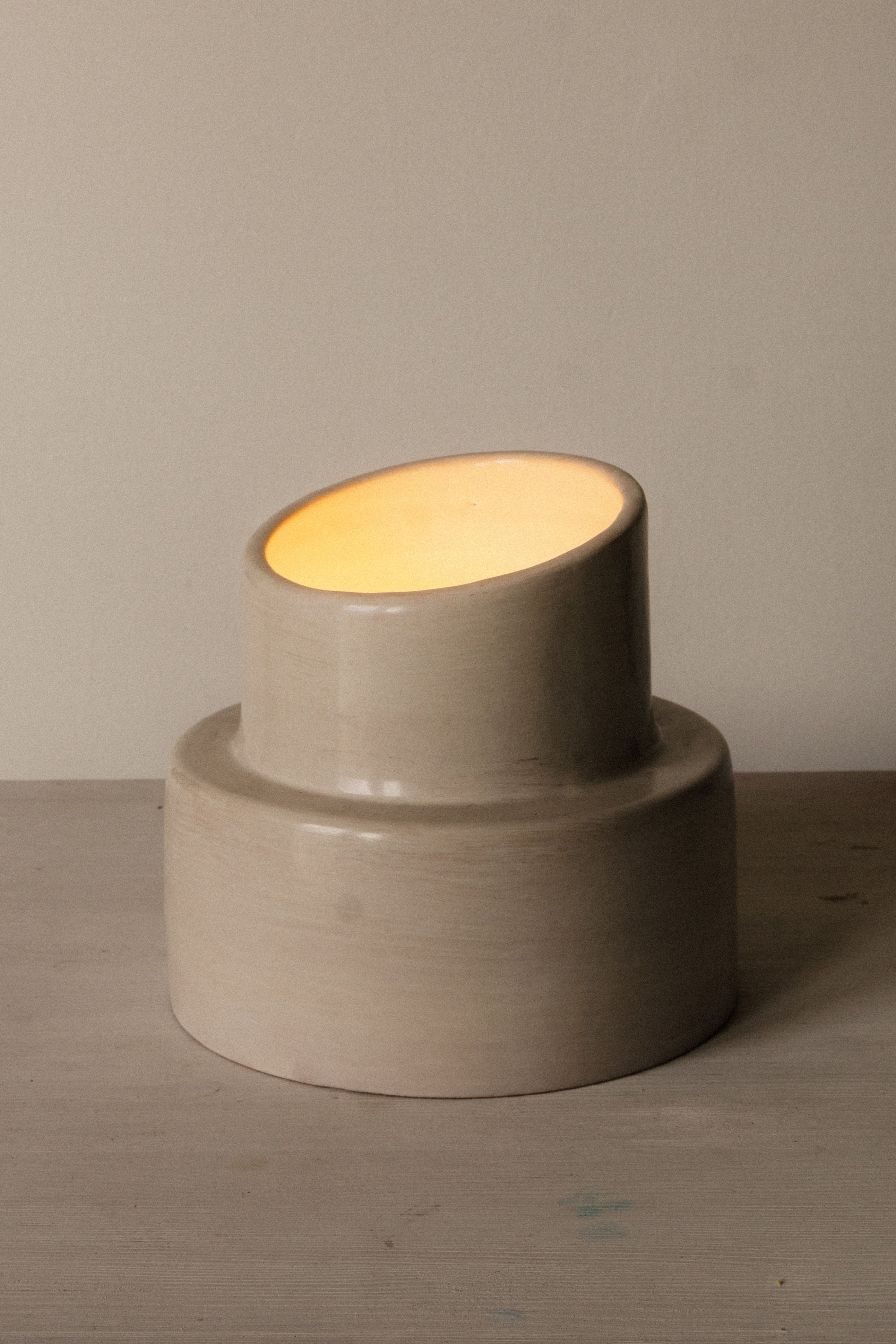 The "Pillar" Table Lamp by COSEINCORSO is a modern beige ceramic lamp with a slanted cylindrical top, casting warm light on a light surface. Its elegant silhouette evokes an ancient column, complementing any neutral-toned space.