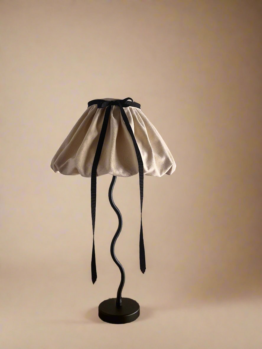 The "Puffball" Table Lamp by Maud Vaughan features a wavy black metal stand and a beige puffball shade crafted from a cotton-linen mix, adorned with a black ribbon. This elegant piece rests on a wooden surface against a plain wall and is hand-sewn in the UK to ensure superior craftsmanship.