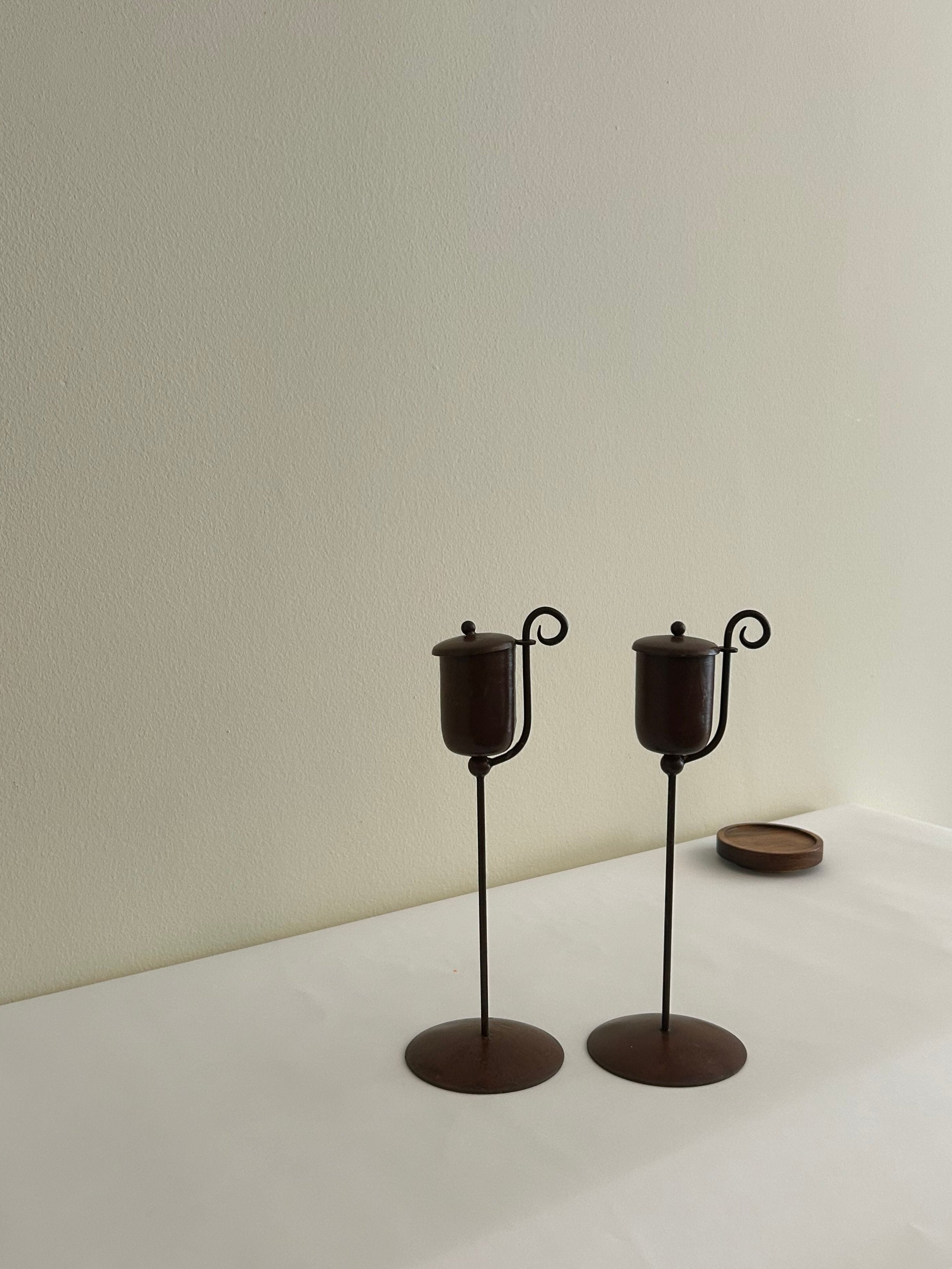 Two elegant, tall candle holders from Flock Studio, crafted in brown metal with curved handles, exude a sculptural aesthetic as they stand on a light surface against a plain cream wall. The Vintage Brutalist Candle Holders have a small, round object slightly visible in the background on the right, adding a touch of intrigue to their design.