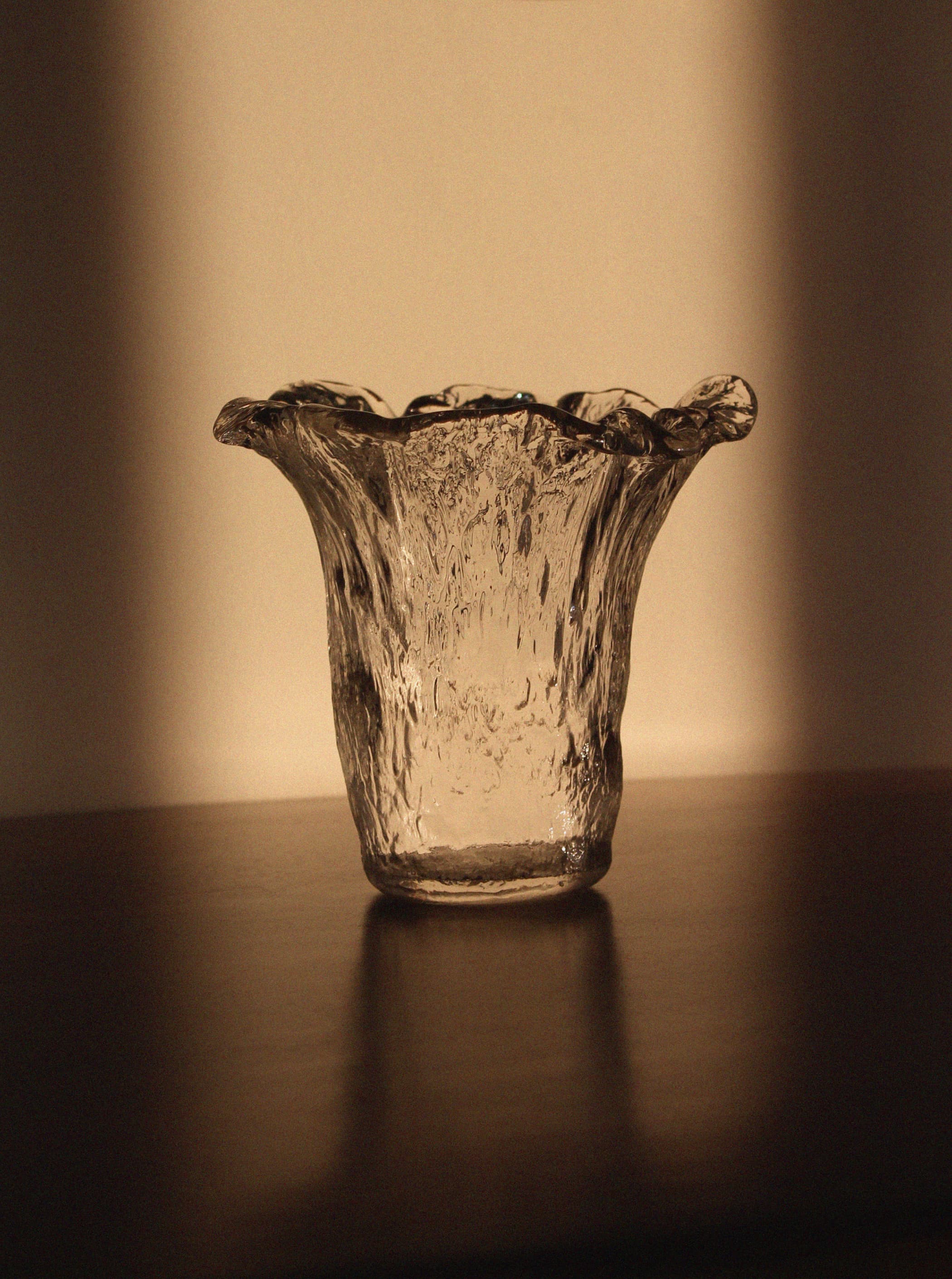 A Uno Westerberg 60s Organic Glass Vase by Vintage Stories, featuring clear textured glass and a flared wavy rim, sits on a dark surface illuminated by warm light that casts gentle shadows in the background.