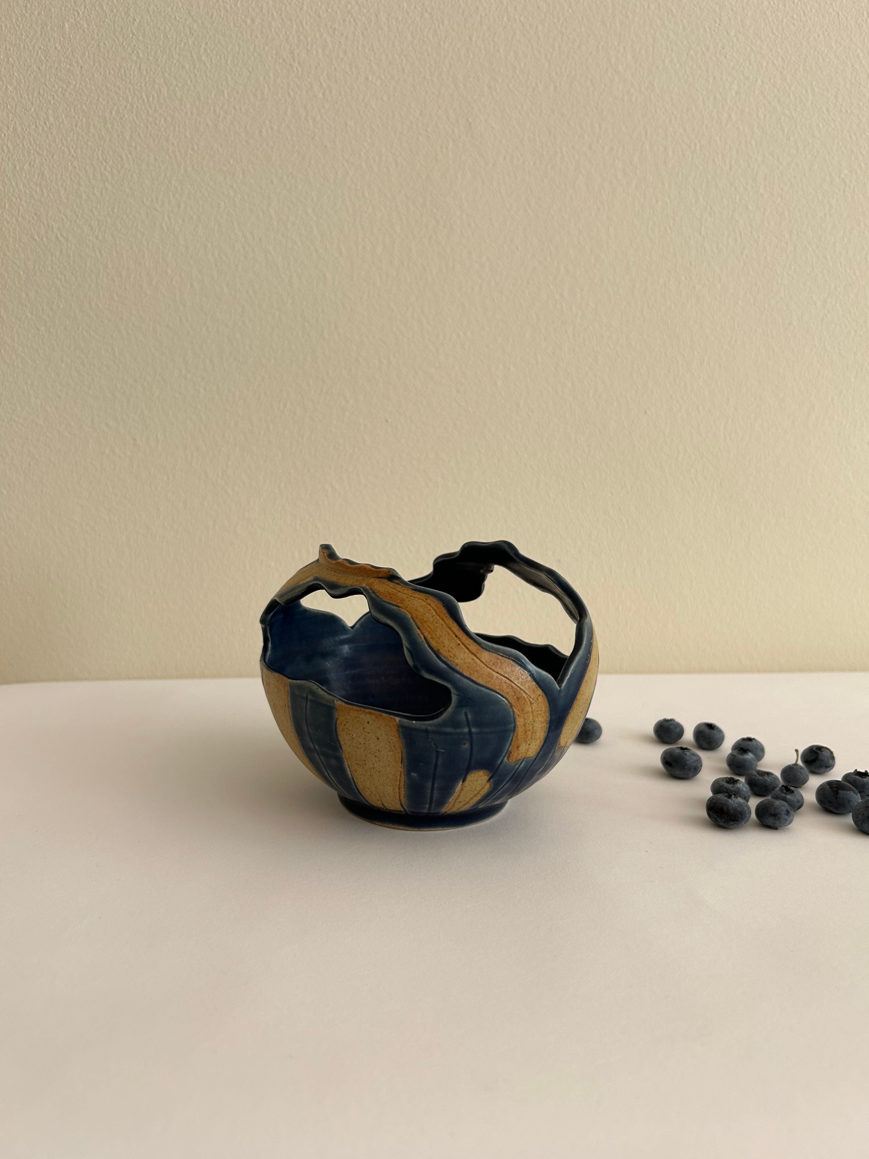 The Artisan Ceramic Bowl by Flock Studio, a small handmade piece with a basket-like shape, showcases blue and brown swirls as it rests on a white surface. Scattered blueberries nearby add a natural touch against the plain cream-colored background.
