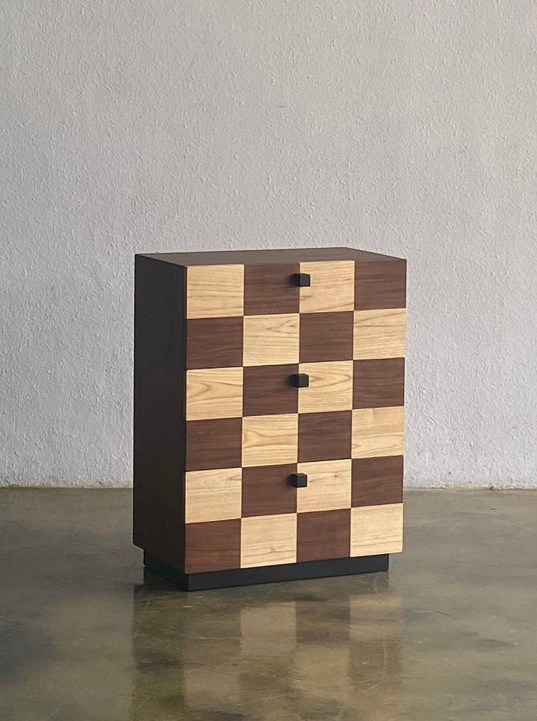 A Coklat Drawer by Studio Kallang with a checkerboard pattern alternating between dark and light wood panels. The extra-wide chest has four storage drawers, each with a small black handle in the center, and it is placed against a plain, light-colored wall on a polished floor.