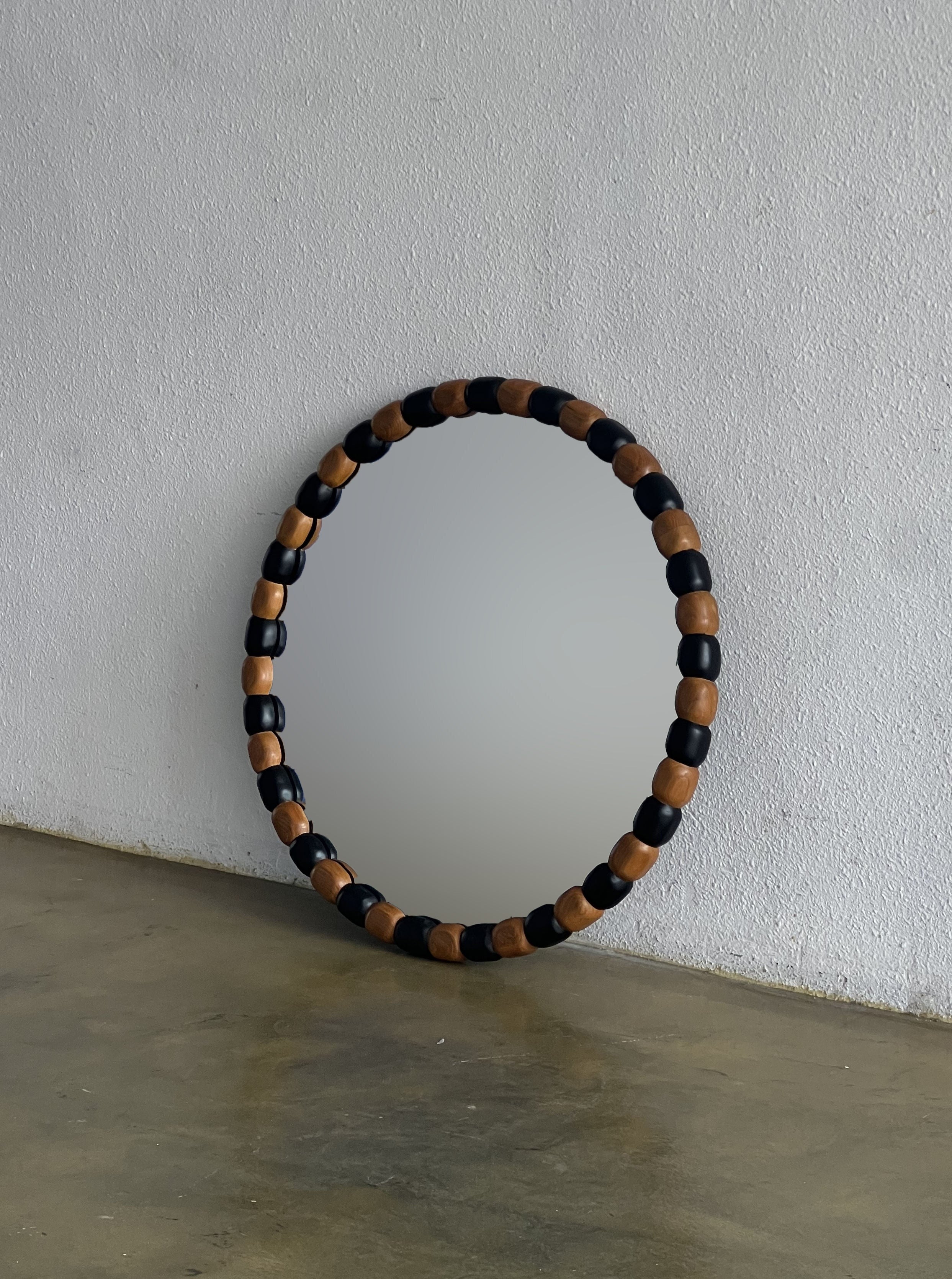 A handcrafted round mirror with a unique frame composed of alternating wood and black segments rests against a plain white wall. The Onde Mirror Round (Teak/Black) by Studio Kallang is placed on a polished concrete floor, creating a minimalistic and modern aesthetic.
