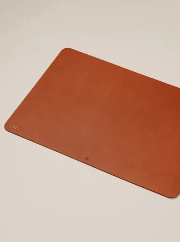 The Paradise Row Leather Desk Mat, featuring rounded corners and subtle "LW" initials embossed in the bottom left corner, is a perfect personalized gift. This rectangular brown leather accessory stands out beautifully on a light surface.