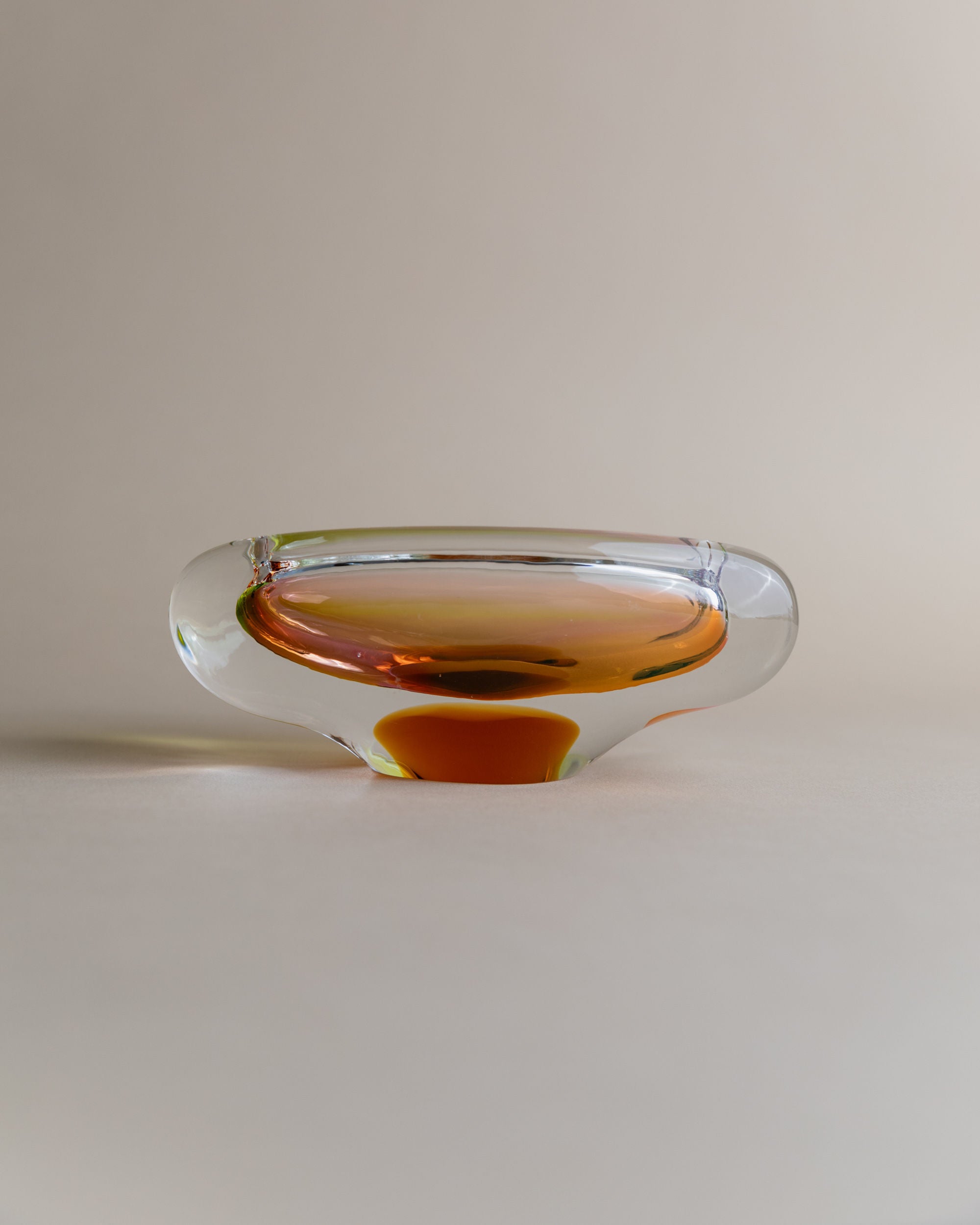 The Glass ashtray by Josef Rozinek for Novy Bor, from Bottega Jacobs, has a unique rounded design with an amber interior. Its transparent material reflects light, creating a gradient effect on the neutral surface it sits on.