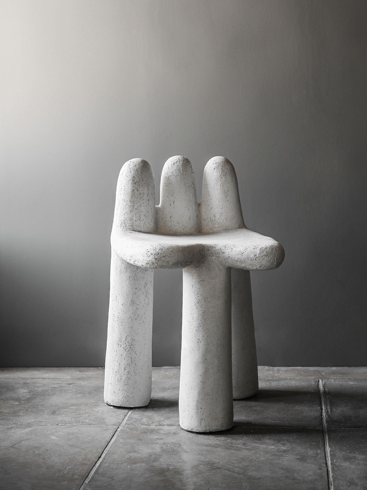 The Washed Concrete Chair by Studio OSKLO features a white, textured finish with three rounded legs and an abstract, organic form. Handmade from molded concrete, this unique piece stands out against a minimalist gray background.