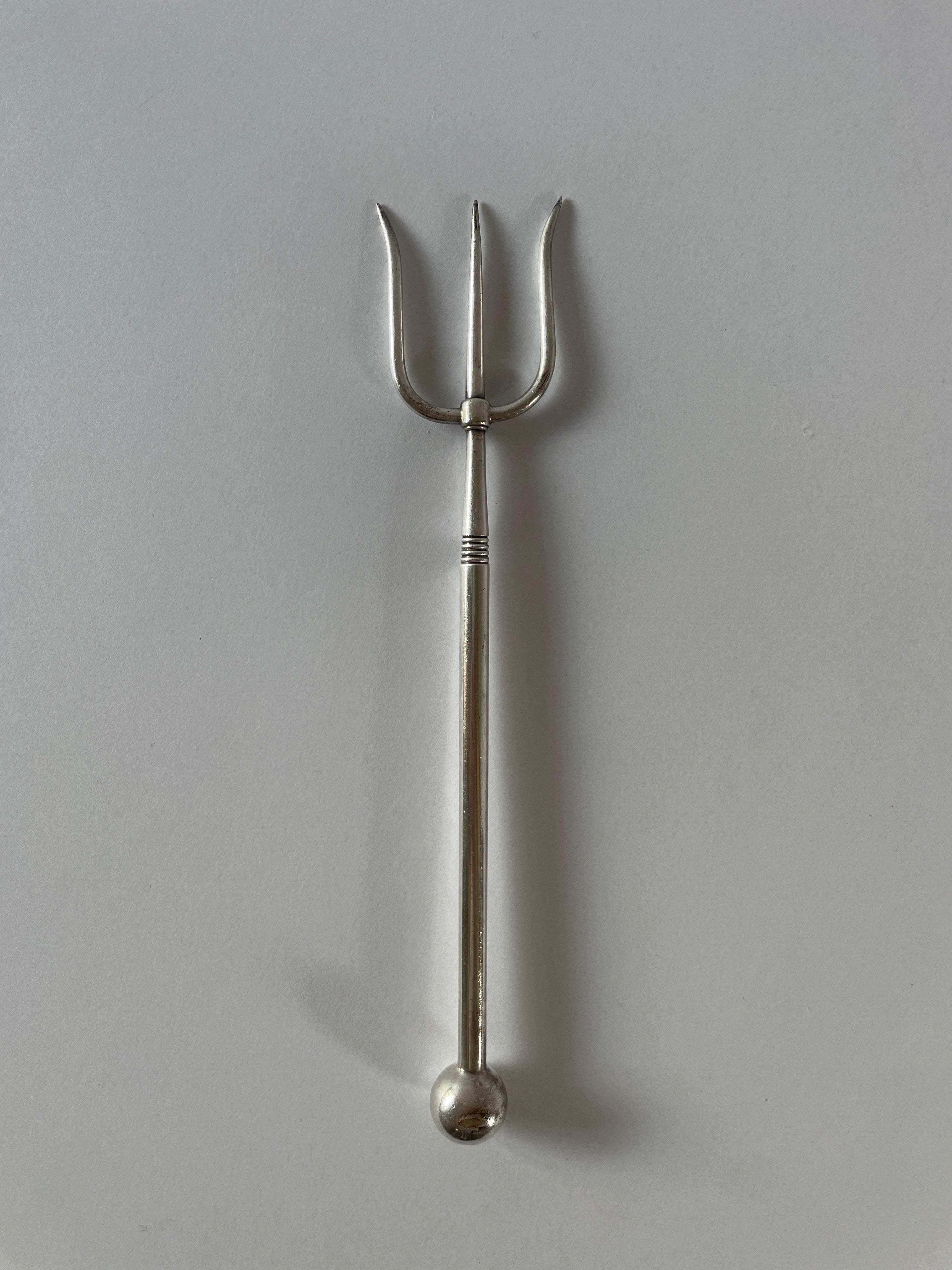 An **Antique Silver Plate Serving Fork** by **Maud Vaughan**, shaped like a trident, rests on a plain light gray surface. This utensil has three curved prongs and a long, smooth handle ending in a rounded sphere—evoking Victorian etiquette.