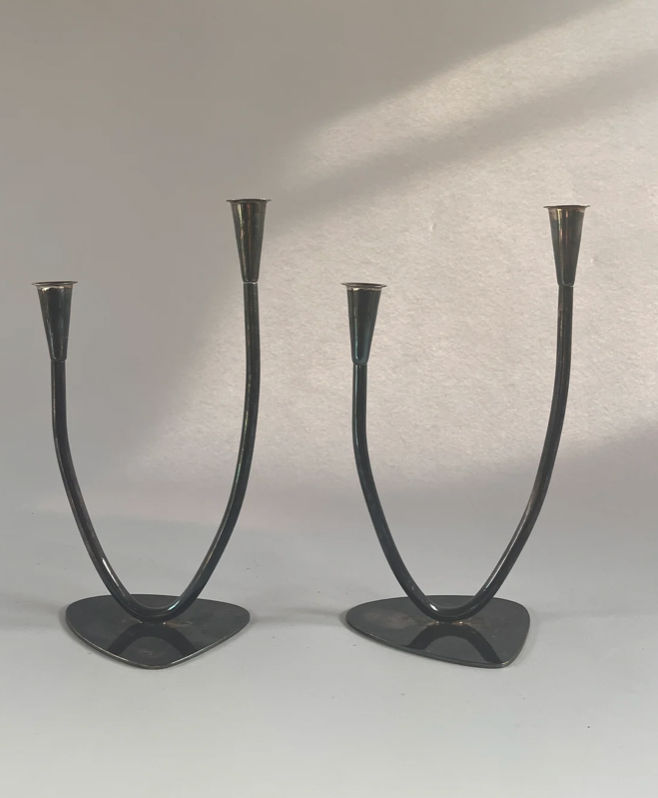 These sleek, modern black candleholders from Maud Vaughan, named the Pair of Mid-century Candleholders, feature triangular bases and curved arms, each designed to hold two candles. Crafted in Germany, they cast soft shadows on a plain gray background.