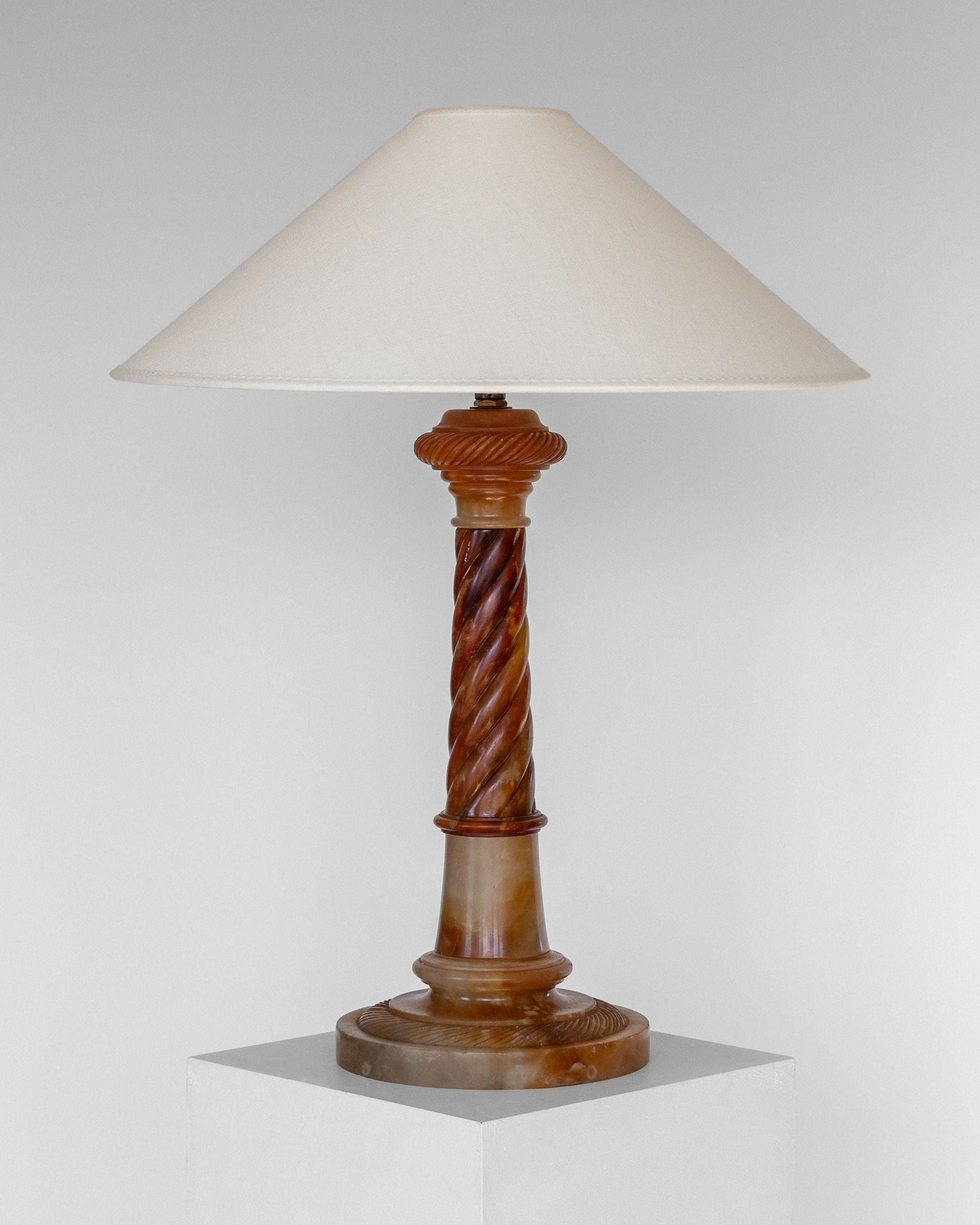 The Antique Alabaster Table Lamp, C. 1900 by Spigel features a beige, conical lampshade and a decorative, hand-carved alabaster base. Displayed against a plain, light gray background and standing on a white pedestal, this lamp boasts a classic and timeless design with an elegant touch that evokes the charm of an antique table lamp.