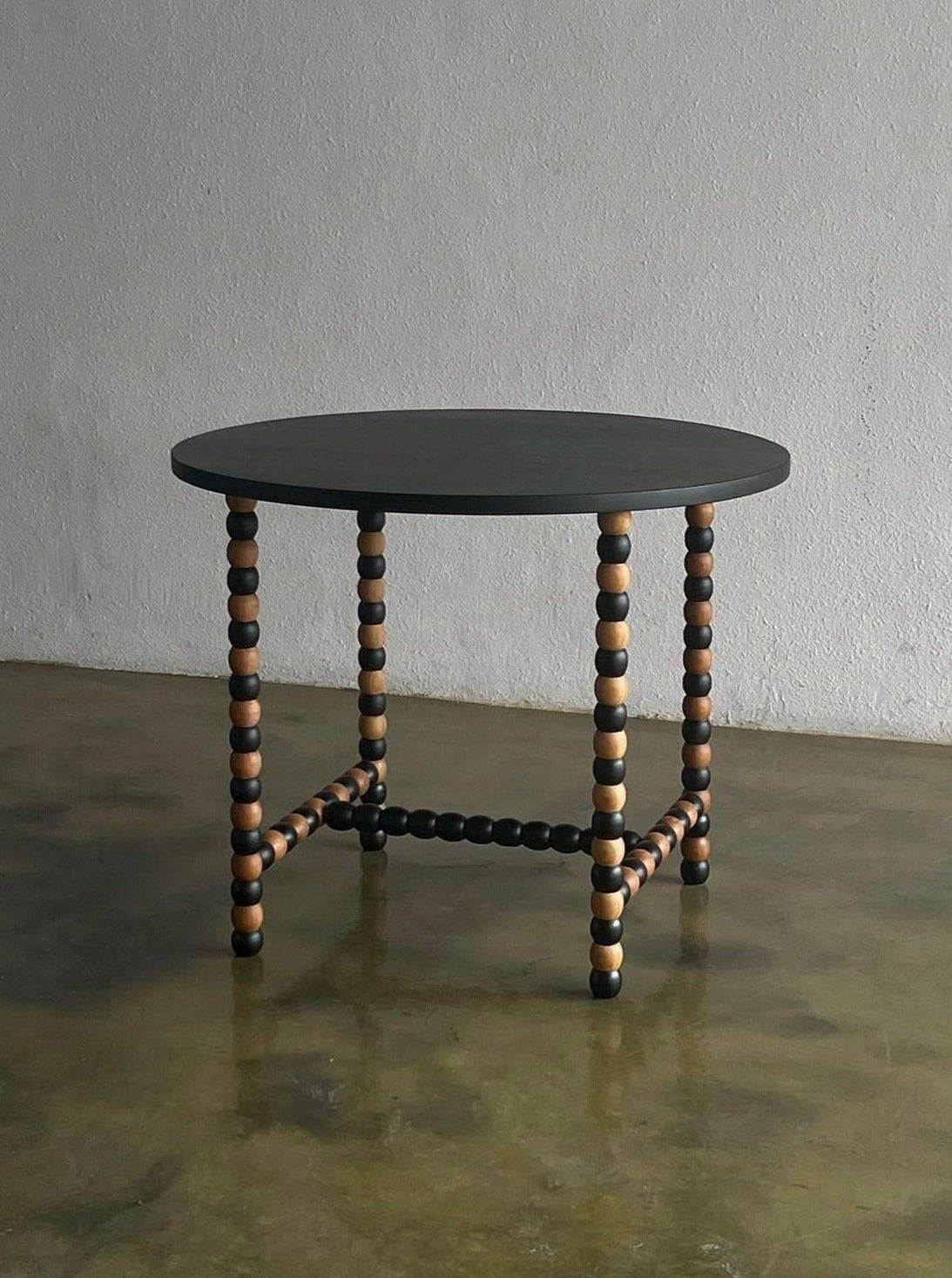A round table with an all-black tabletop stands on four legs made of alternating black and natural wood-colored beads. This two-toned wood table, the Onde Round Table by Studio Kallang, is positioned on a concrete floor against a plain grey wall.