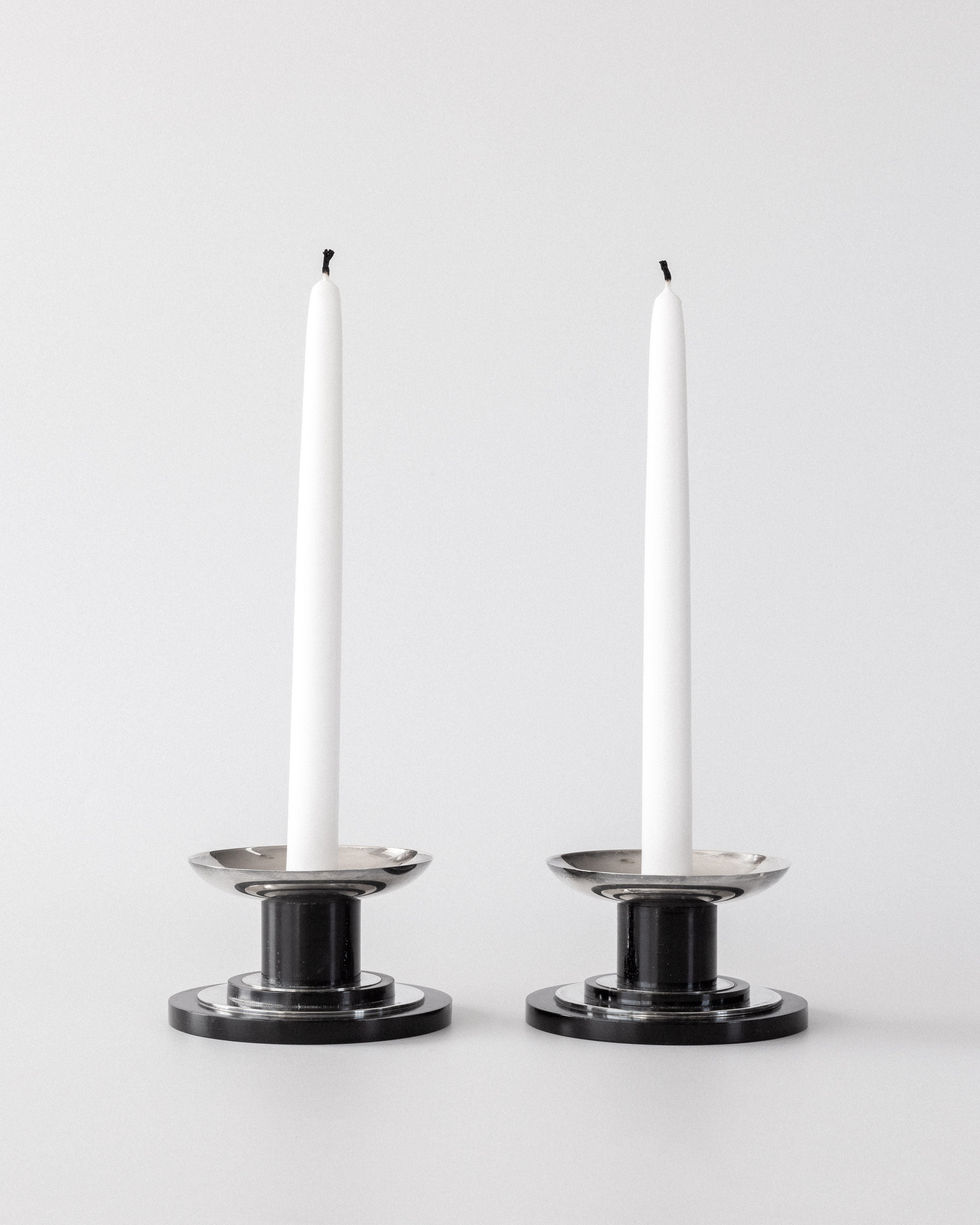 Two white taper candles stand upright and unlit in Spigel’s Modernist Sterling Silver Candleholders (1934), set side by side against a plain light gray background.