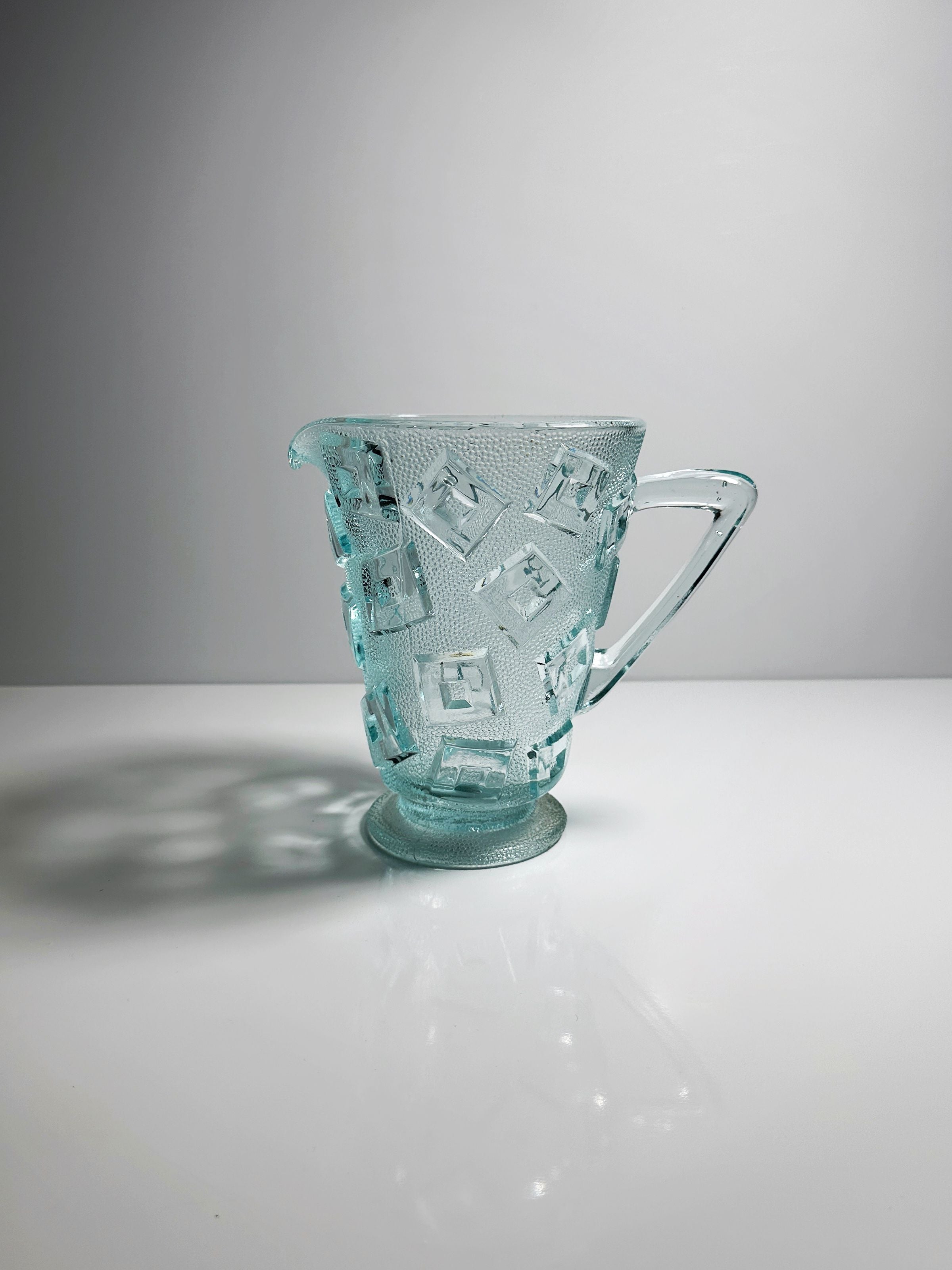 The Bohemian Art Deco Carafe 30s by Dodo Vintage, made from translucent pressed glass, features a textured pattern of raised geometric shapes. It rests on a reflective white surface against a light gray background, showcasing its cubist style. The carafe casts a soft shadow to the side with its handle and spout.