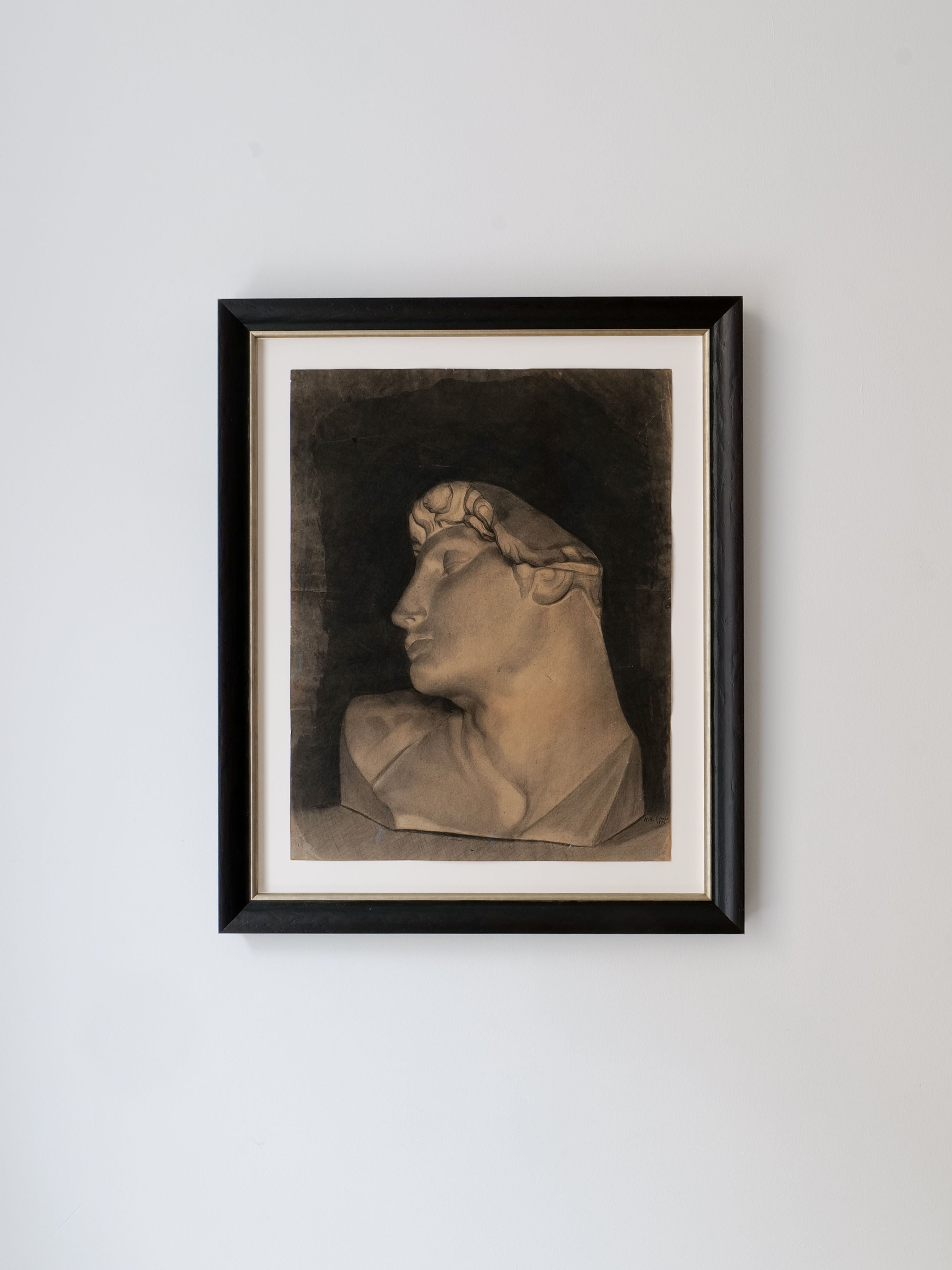 The "Charcoal Sketch 1894" by Collection apart is displayed on a light gray wall, resembling a Michelangelo sculpture with its classical-style bust in profile. This artwork in sepia and black emphasizes the serene expression and intricate details. The piece is elegantly framed with a black frame and white border, reminiscent of those found in the Louvre Museum.