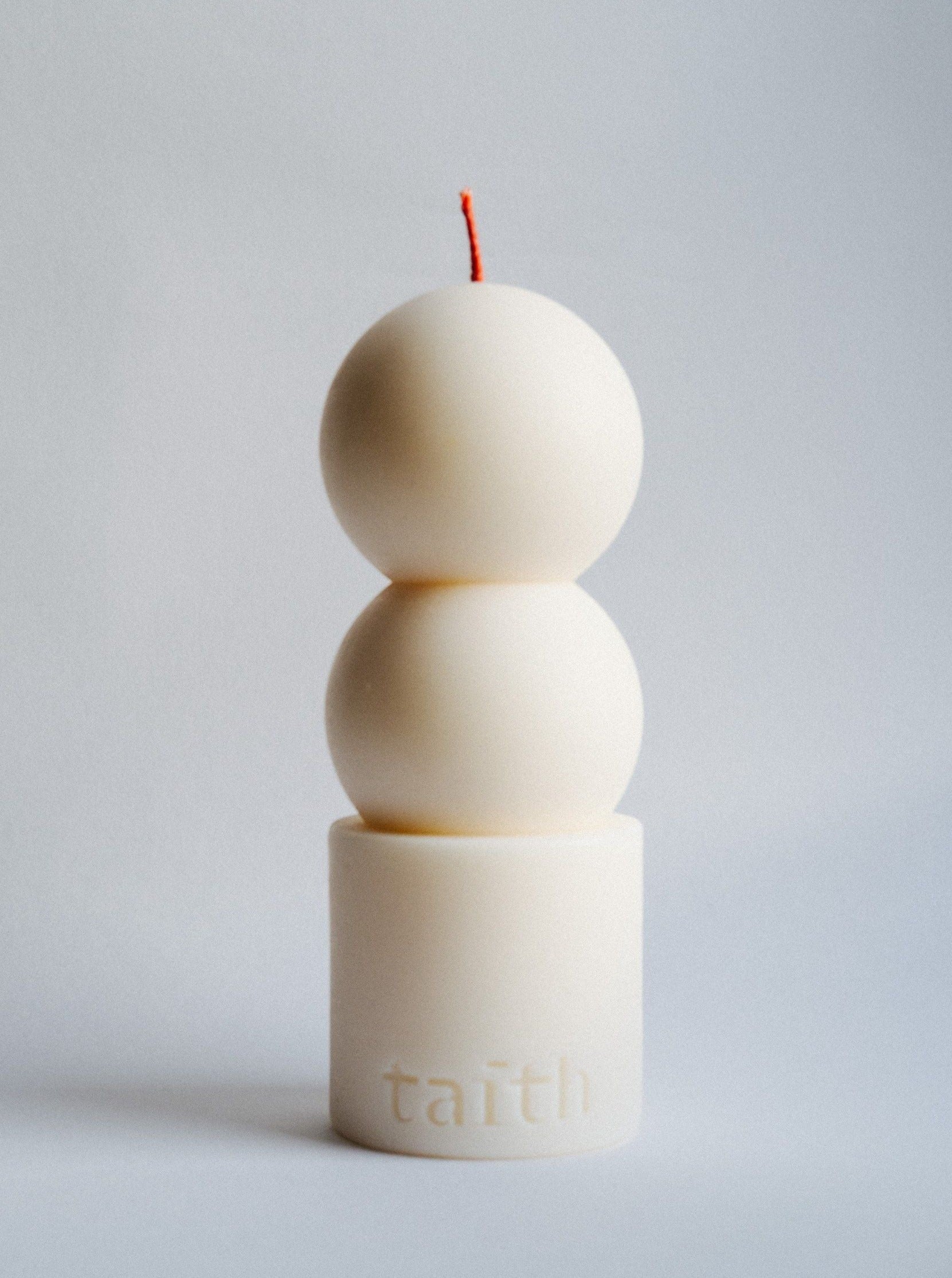 A minimalistic white Lilu Candle Cream crafted in the shape of three stacked spheres atop a cylinder base, with the word "Taith" engraved on the base. The eco-conscious ambiance is accentuated by the natural soy wax and single red wick at the top. The background is plain and light gray.