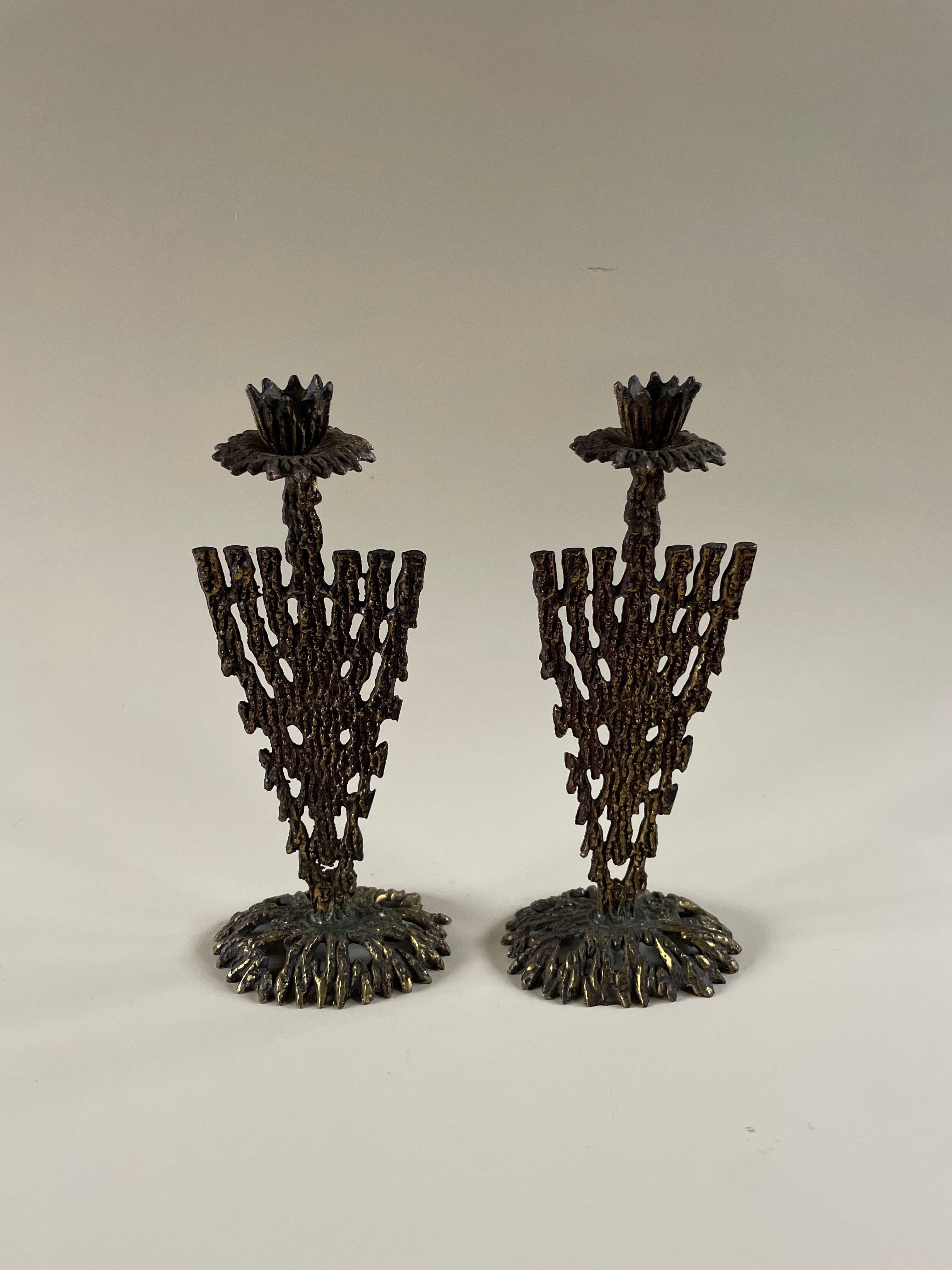 The Pair of Brutalist Metal Candleholders by Maud Vaughan exudes a vintage 1970s charm, showcasing intricate tree-like motifs on a minimalist white backdrop. Each candle holder is adorned with detailed floral patterns at the top, and features a textured base that resembles roots.